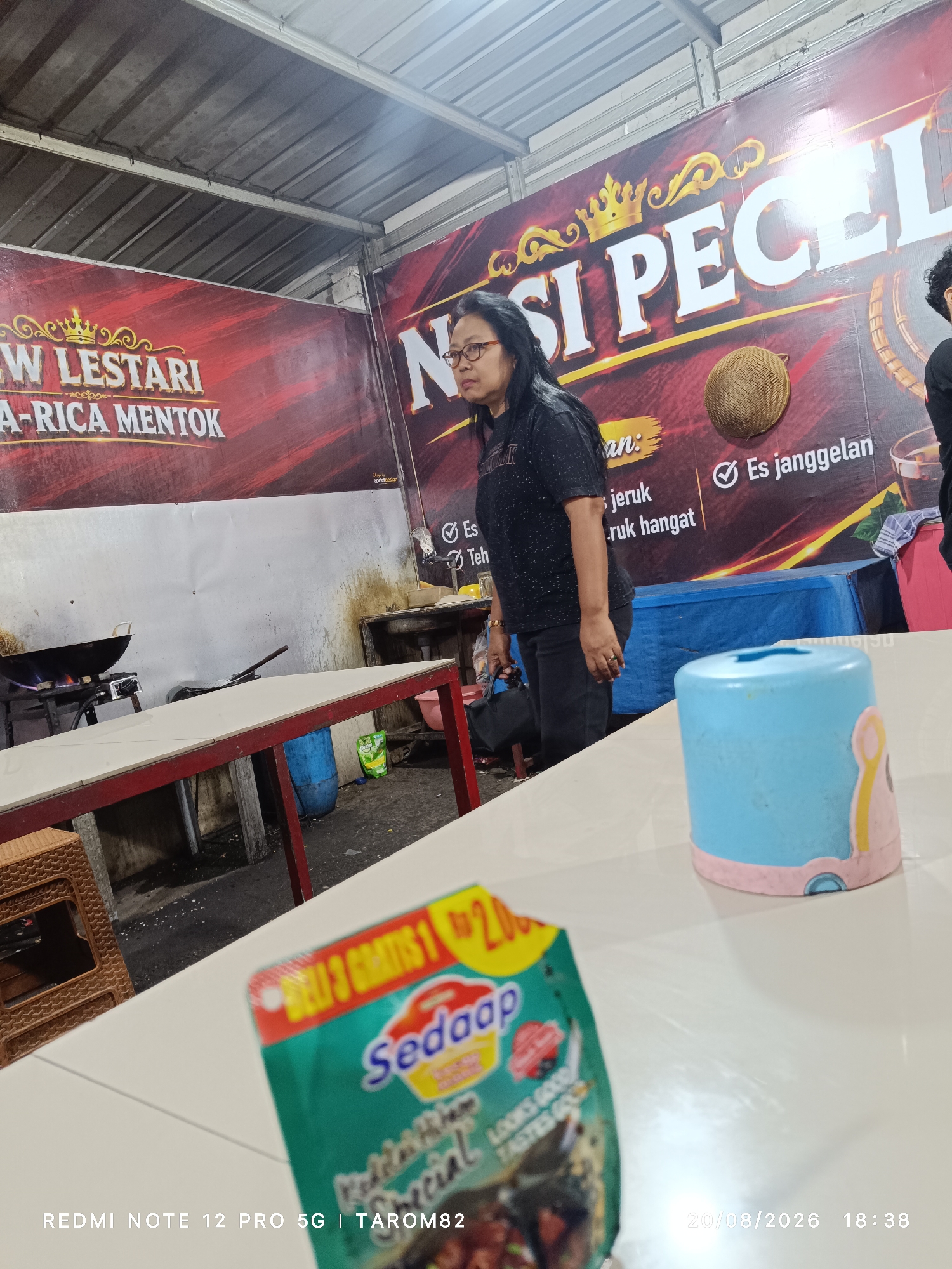 The image depicts an indoor scene at what appears to be a food stall or restaurant in Bringinbendo, Indonesia. In the background, a large banner displays "NASI PECEL" in bold white lettering, suggesting this is the establishment's specialty. To the left of the banner, another banner reads "W LESTARI" and "A-RICA MENTOK". A woman with glasses and dark hair, wearing a black t-shirt and black pants, stands in front of the banner, looking towards the right. She is wearing a ring on her left hand. A dark bag is visible at her feet.
The environment includes tables set up for dining. One table in the foreground is covered with a white tablecloth, and on it rests a blue and pink tissue holder. Another table, further back and with a red frame, has a wok on a burner, with flames visible, indicating food preparation. Various items are scattered around the preparation area, including a blue plastic barrel, a green packet, and what looks like a sink. The ceiling is made of corrugated metal. In the lower right corner of the image, a snack packet with the brand "Sedaap" is partially visible, along with a date and time stamp: "20/08/2026 18:38". Text at the bottom of the image reads "REDMI NOTE 12 PRO 5G | TAROM82".