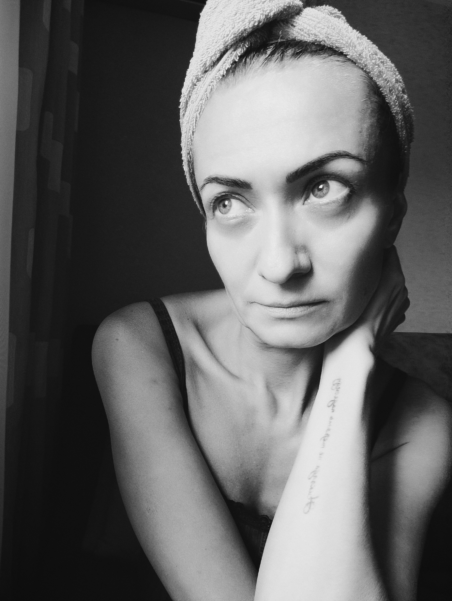 This is a black and white, close-up, profile shot of a woman with a towel wrapped around her head. She is looking to her left with her chin resting on her left hand, which is cupped around her neck. The woman is wearing a dark-colored tank top. A tattoo is visible on her right forearm. The background is dark and appears to be a curtain on the left side and a dark wall on the right. There is no indication of location within the city of Moscow, Russia.
