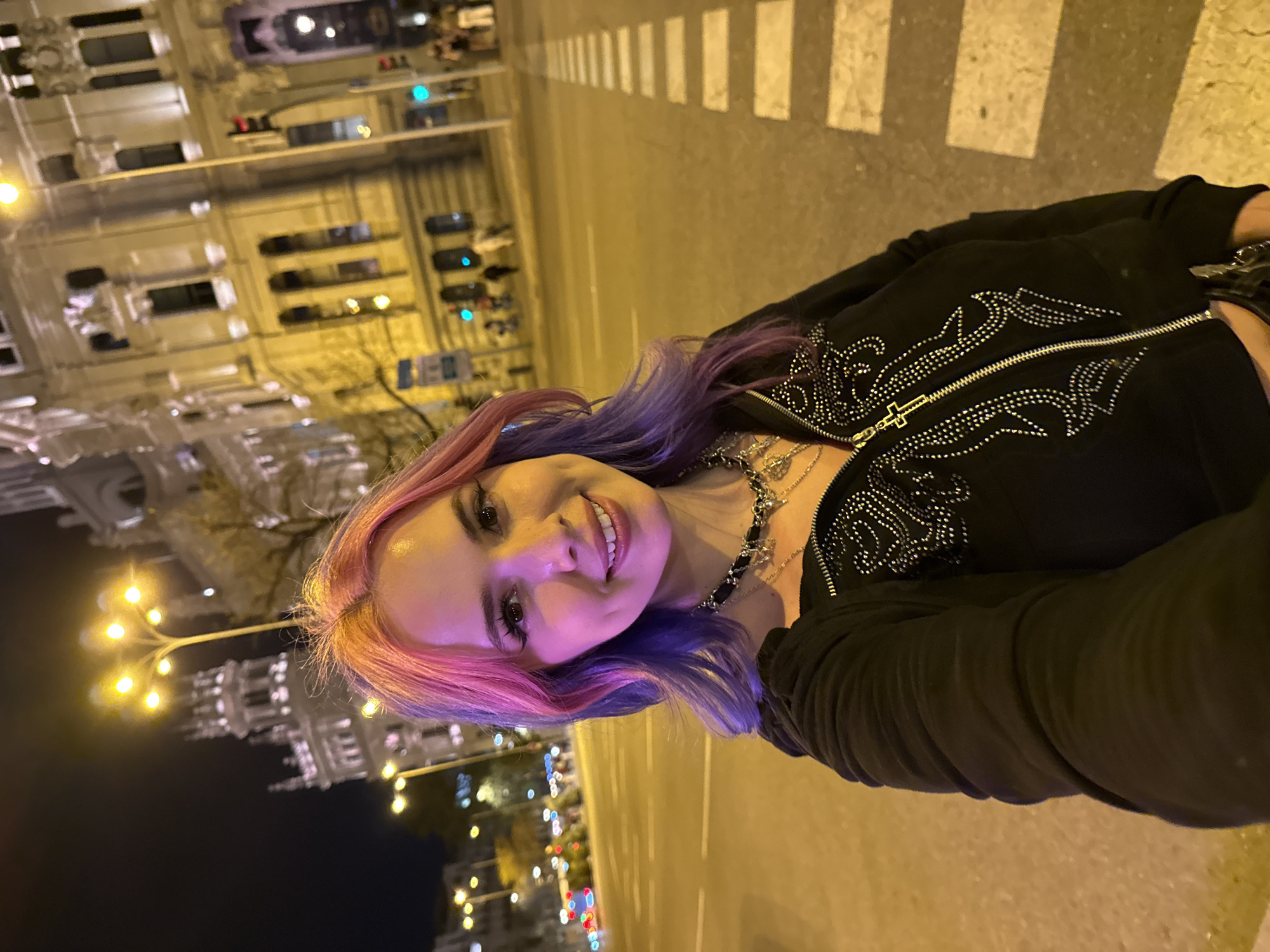 A woman with pink and purple hair is standing in a street at night. Behind her is a large, illuminated building and a street with pedestrian crosswalks and parked cars. The building appears to be the Palacio de Cibeles in Madrid, Spain. The woman is wearing a black jacket with silver embellishments and several necklaces. She is smiling at the camera.