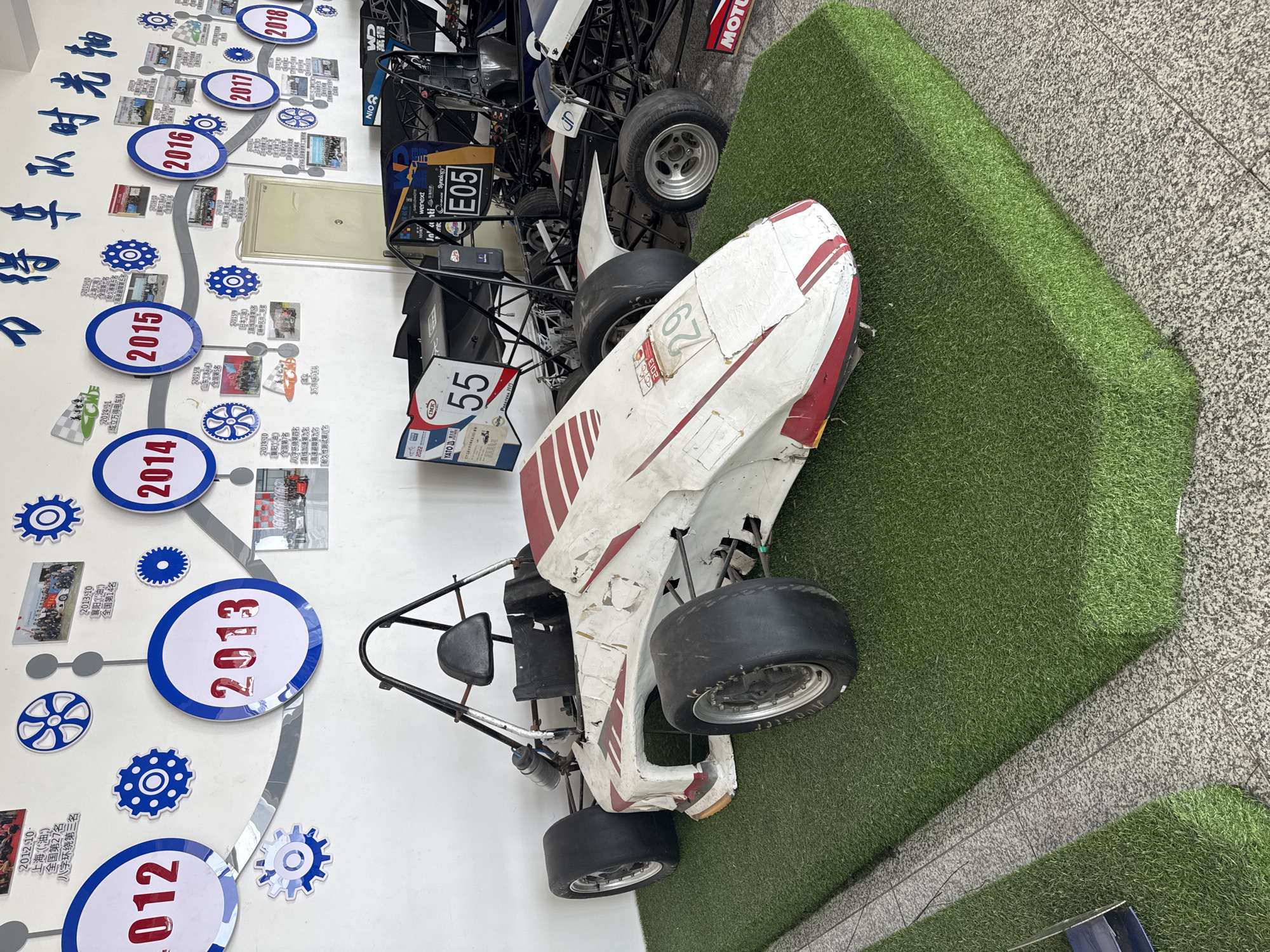 The image captures a scene featuring a go-kart, possibly displayed in an exhibition or a museum-like setting. The kart, predominantly white with red stripes and the number "67" on its side, appears weathered and potentially damaged. It rests on a patch of artificial grass next to a gray tiled floor.
On the wall behind the kart, there is a timeline display. The timeline is decorated with circular markers indicating years from 2012 to 2018. These markers also contain smaller images and text in Chinese. The text in the timeline indicates the various achievements and activities of an electric car team from Wande, China.
To the left of the kart, there's a sign with the number "55" written on it, which is next to another go-kart chassis, this time dark colored and in the background. The image's location is likely in Zhongtun, China, as indicated by the Chinese characters on the timeline display. The environment appears to be indoors.
