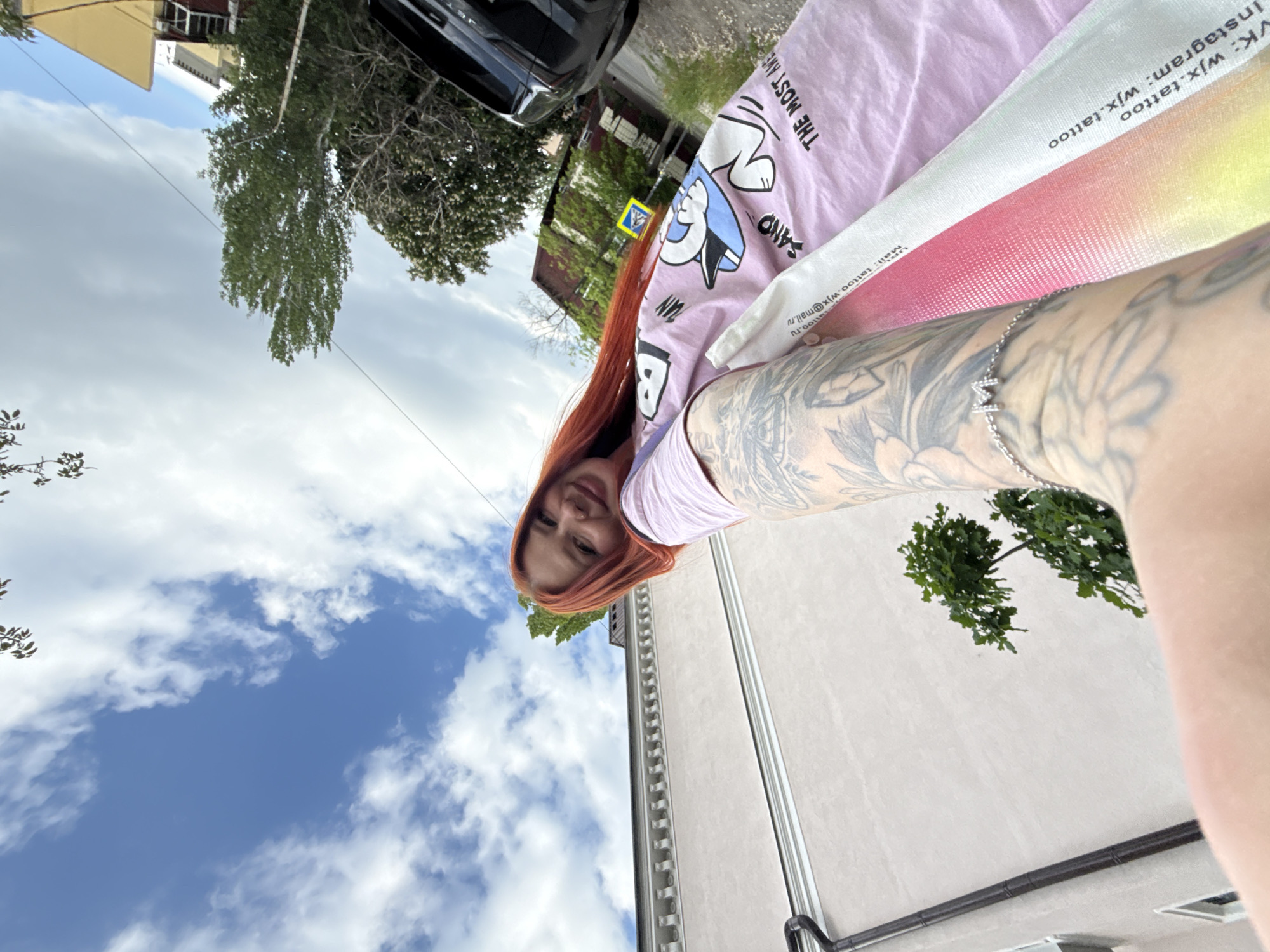 The image shows a woman with red hair and multiple tattoos on her arm taking a selfie outdoors. She is wearing a light purple t-shirt with cartoon illustrations. She is holding a promotional paper that contains the information "VK: wjx.tattoo Instagram: wjx.tattoo wall: tattoo.wjx@mail.ru", indicating it may be related to a tattoo artist or studio.
In the background, there is a blue sky with some clouds, a building, trees, and part of a black vehicle. Based on the content visible in the promotional paper, it's likely the photograph was taken in Rostov-on-Don, Russia.