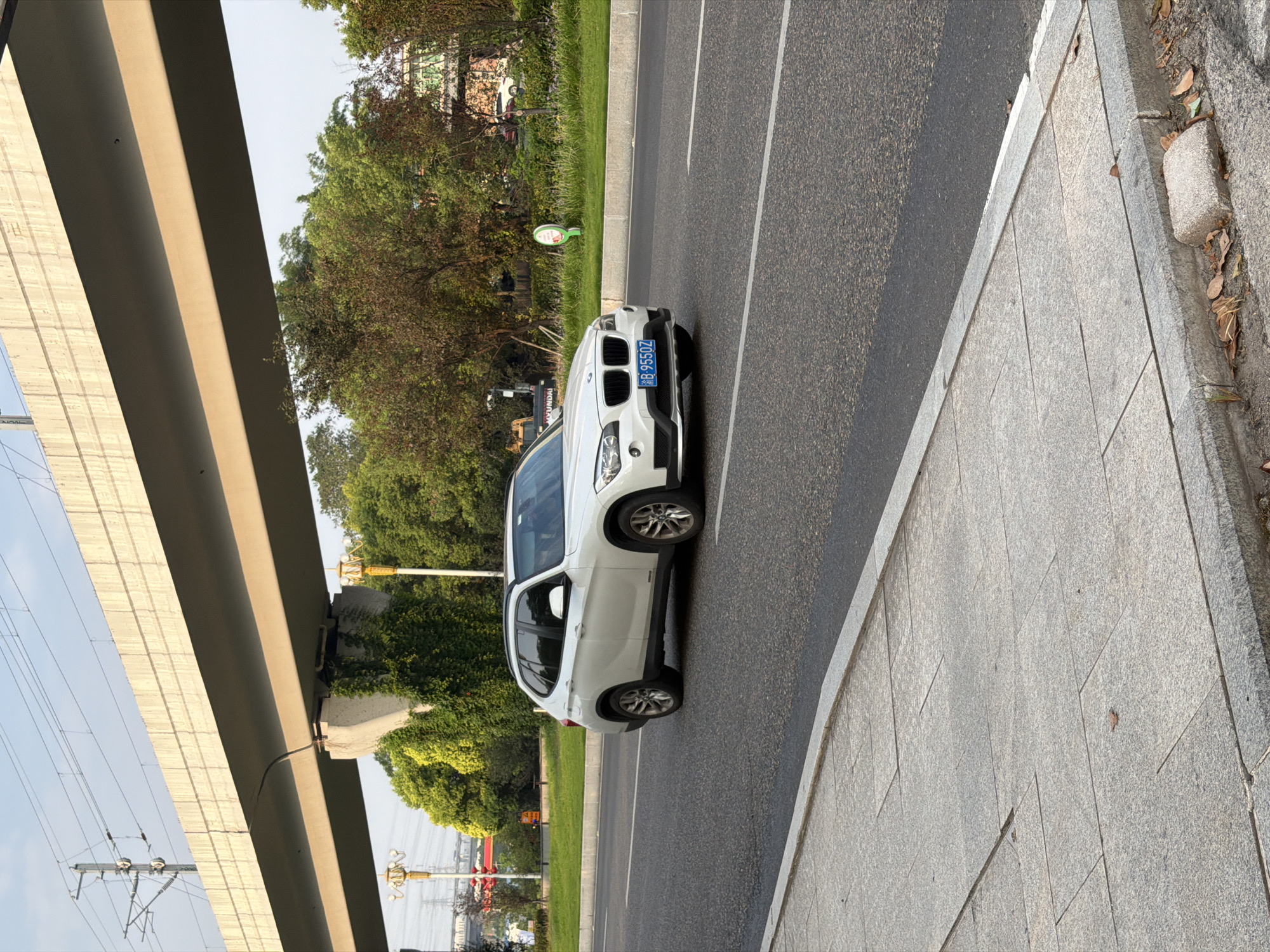 A white BMW X1 SUV, license plate B-9550Z, is driving on a multi-lane paved road in Jiangjin, China. The vehicle is positioned in the right lane, traveling away from the viewer. The road has a gray asphalt surface with white lane markings. To the left of the road, there is a concrete sidewalk with large, square gray tiles. A concrete curb separates the road from the sidewalk.
An elevated concrete overpass, likely part of a transportation infrastructure such as a highway or railway, spans across the upper portion of the image. The overpass is supported by concrete pillars, one of which is partially covered in green ivy. Overhead power lines and their supporting structures are visible above the overpass.
The background consists of a grassy median strip with some trees and greenery. In the distance, there are more trees and some buildings, including a glimpse of a taller structure with a green facade on the far right. The sky is clear with a pale blue hue, indicating daylight. The overall scene appears to be an urban or semi-urban environment, possibly a road leading into or out of the city. There are no people visible in the image. The lighting suggests it is daytime, likely late afternoon due to the angle of the sun casting shadows.