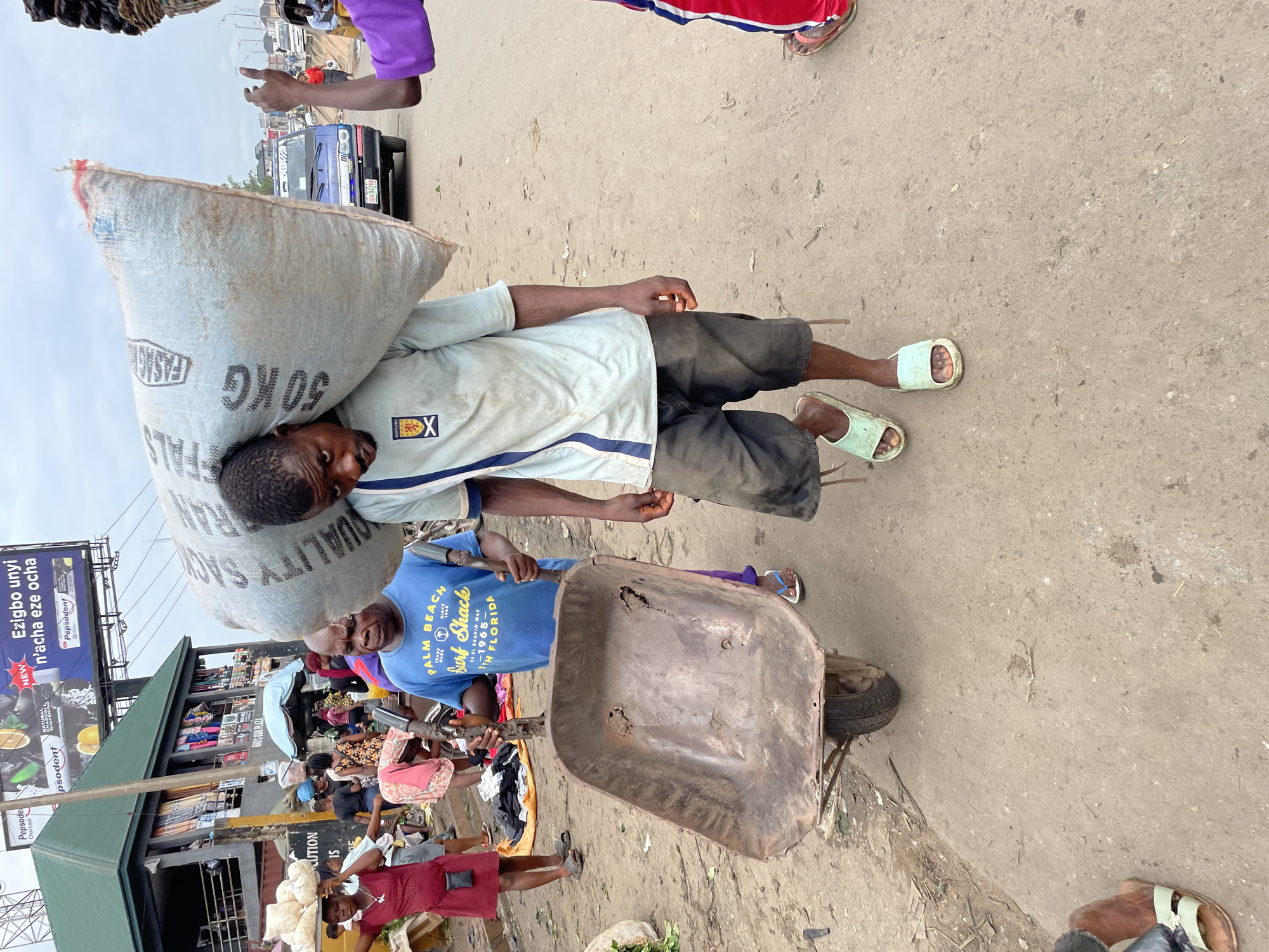 A man carrying a large sack on his shoulders walks down a dirt road in Onitsha, Nigeria. The sack is light blue with visible lettering. He is wearing a light blue t-shirt and dark shorts, and light green flip-flops. To his left, another man in a blue t-shirt and dark pants walks beside a rusty wheelbarrow. In the background, people are gathered around market stalls, with a large billboard visible above a building. A blue van and other vehicles are also present on the road. The scene depicts a busy marketplace environment in the city.