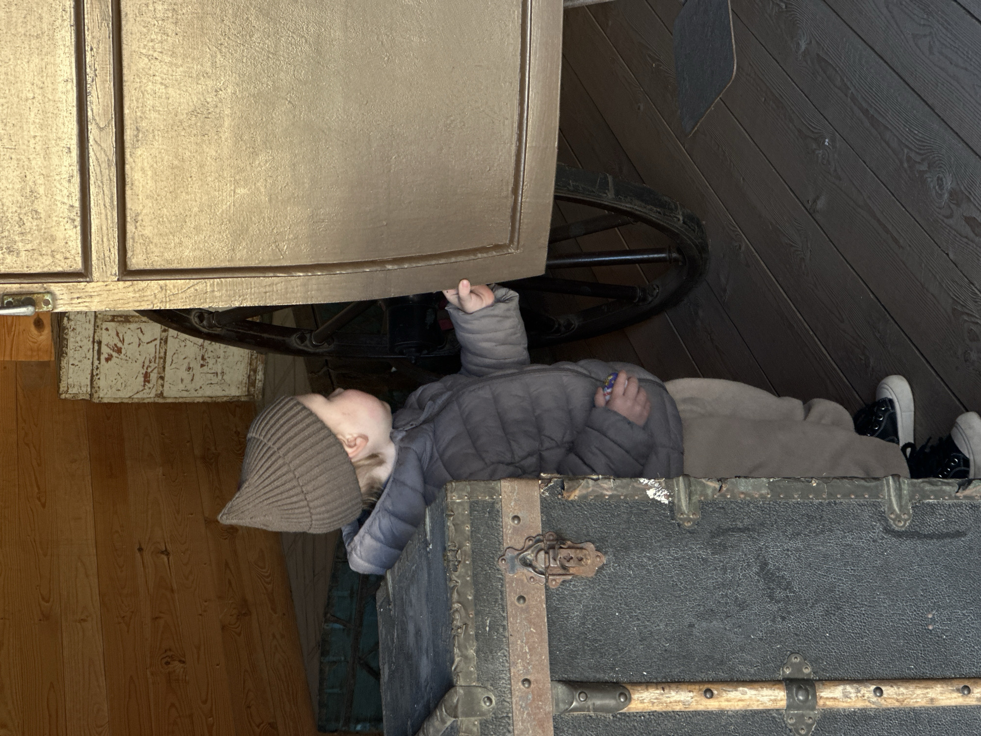 A young child wearing a gray knit hat and a gray puffer jacket lies on an old black trunk with metal trim. The child is partially under a large, gold-colored cabinet with a large wagon wheel visible above them. The child's feet, clad in black and white sneakers, point towards a dark wooden wall in the background. The scene is indoors, with wooden flooring visible on either side of the trunk. There is no verifiable information in the image to indicate it is located in Votkinsk, Russia.