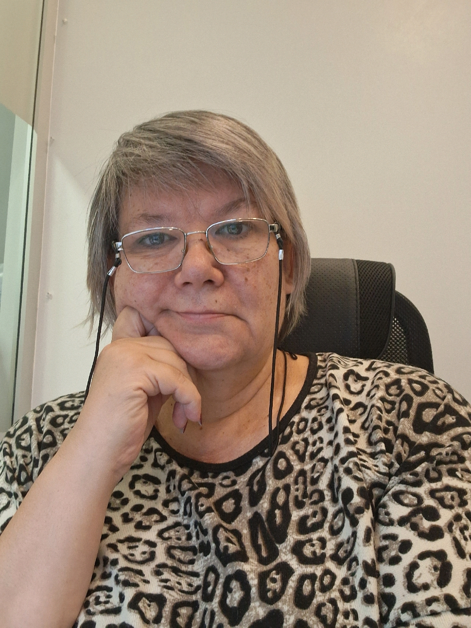 A medium shot captures a woman from the chest up, seated in an office chair. She has short, graying hair and wears silver-rimmed glasses. Her left hand is raised, with her chin resting on her knuckles. She is dressed in a tan and black leopard-print top. A thin black cord, attached to her glasses, hangs down the right side of her face. The background is a plain, light-colored wall, and to the left, a sliver of a window frame is visible. There is no visible indication of the city of Novosibirsk, Russia in this image.