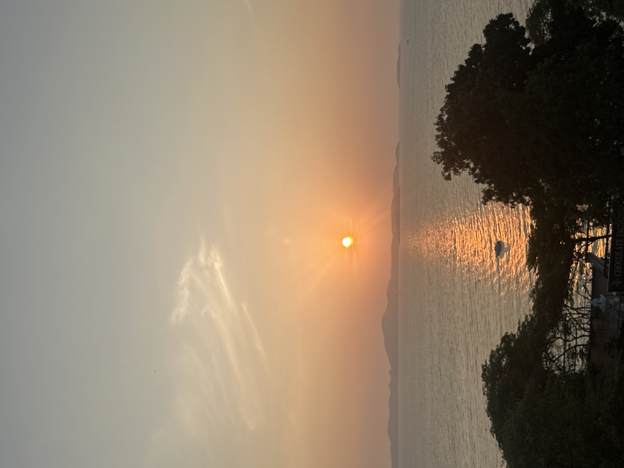 The image captures a sunset over the ocean in Pattaya City, Thailand. The sun is positioned near the center of the frame, casting a bright reflection on the water's surface. The sky is a gradient of colors, ranging from light gray to orange near the sun. A wispy cloud formation is visible in the left portion of the sky. The ocean appears calm, with a few distant landmasses visible on the horizon. Trees occupy the right side of the frame. A small boat is visible on the water. A sign with the text "CHOCOLATE FACTORY" is visible on the right of the scene.