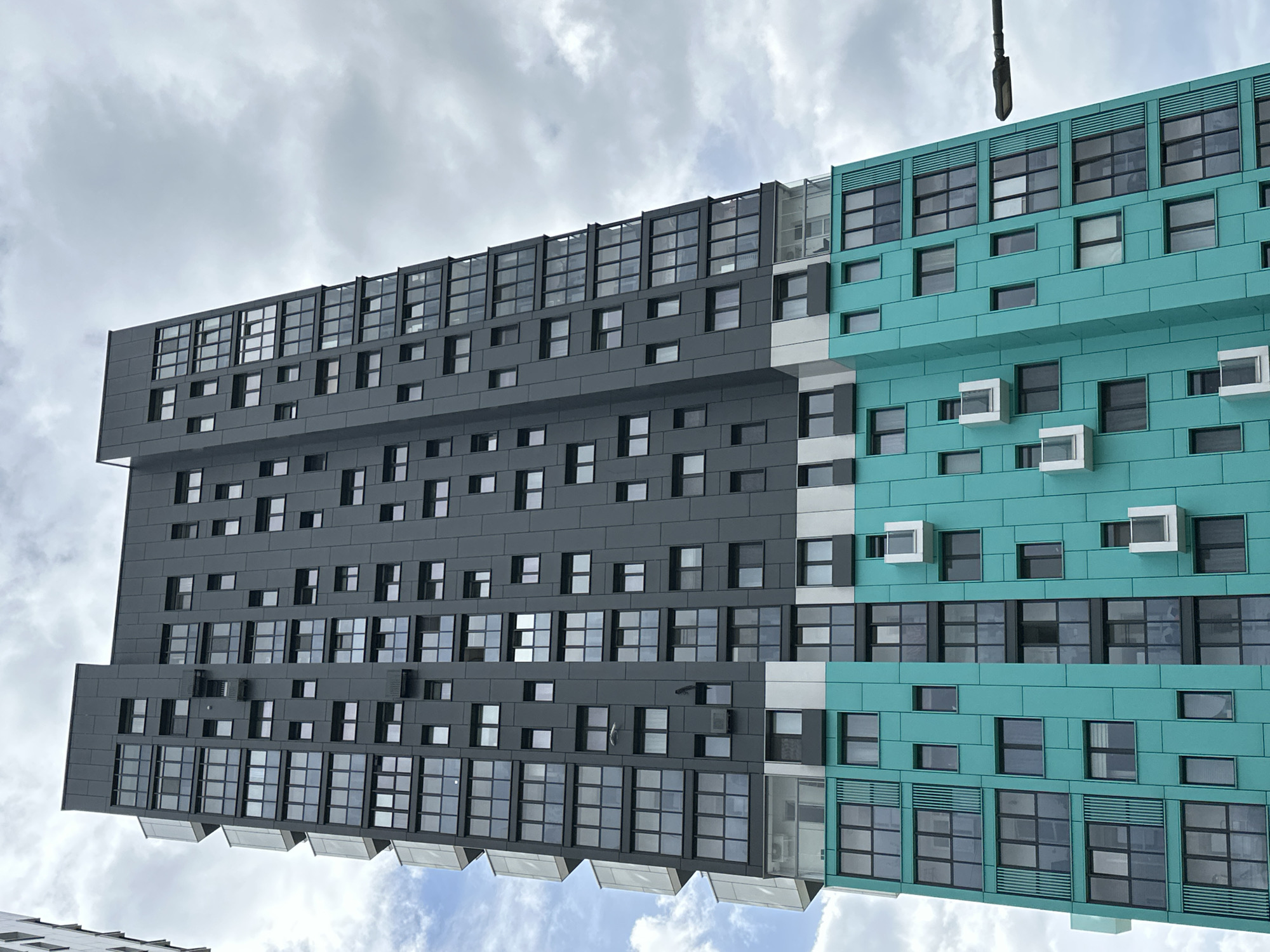 A low-angle, slightly rotated photograph shows a modern building in Бараўлянскі сельскі Савет, Belarus. The building is divided into two distinct color sections: the left side is gray, and the right side is teal. Both sides have numerous windows of varying sizes and configurations, creating a grid-like pattern across the facade. The teal side of the building features protruding, box-shaped elements around some of the windows. At the very top, both sides have a row of large windows covered with some kind of window cover. The sky is overcast with diffused, white-gray clouds filling the upper portion of the frame, with hints of blue sky visible in the bottom section. An object resembling a light pole is visible at the top of the image.