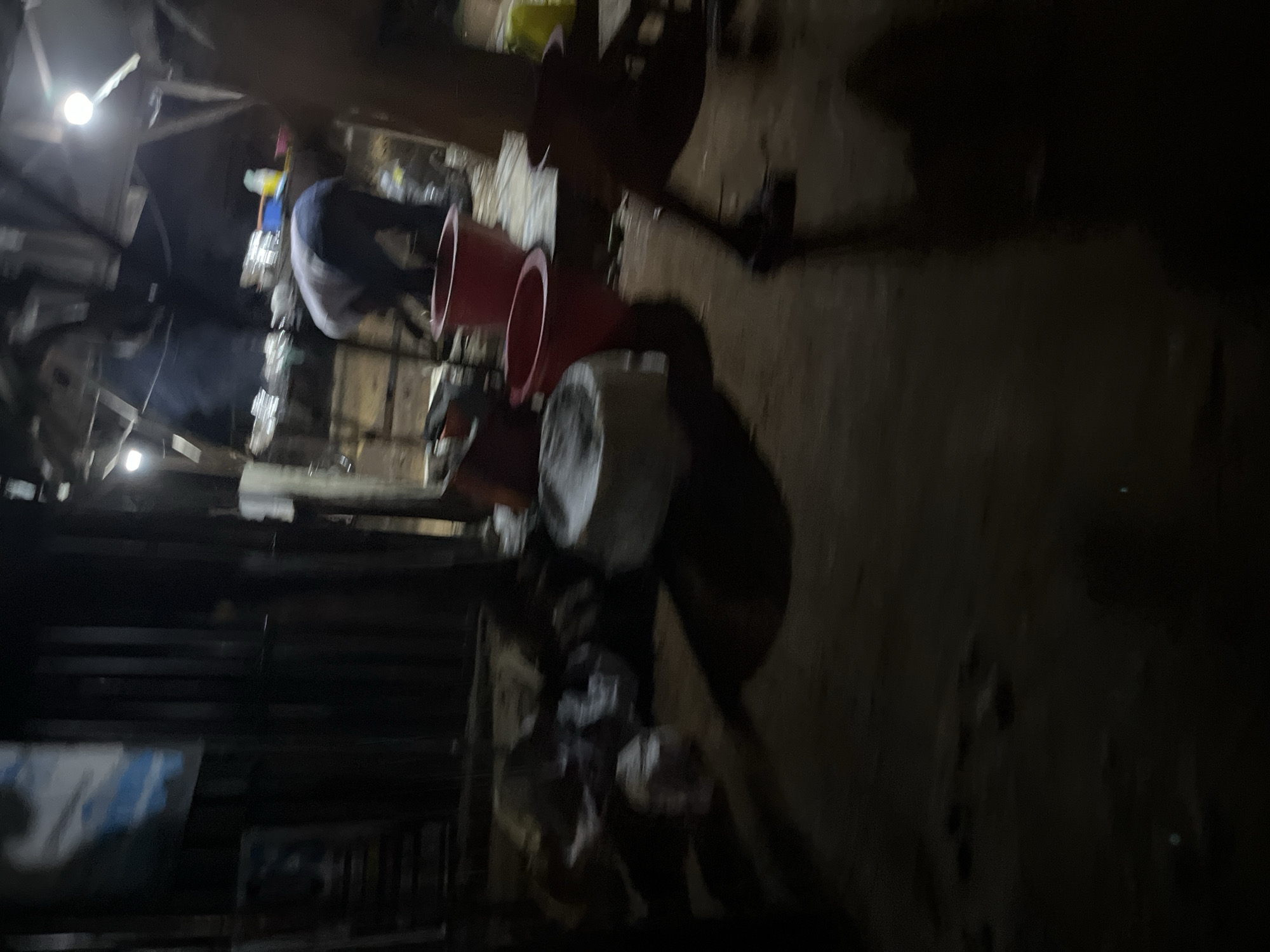 The image depicts an outdoor scene at night, possibly in a market or food stall area, with dim lighting provided by overhead bulbs. In the background, a person wearing a light-colored shirt and dark pants is bent over, appearing to be engaged in some activity. Several large red buckets and a white container are visible in the foreground and midground. There is steam or smoke rising in the background, suggesting cooking or heating is occurring. The ground appears to be dirt or sand, and there are various bags and debris scattered around. The overall impression is one of a busy, functional, and somewhat cluttered outdoor workspace.
