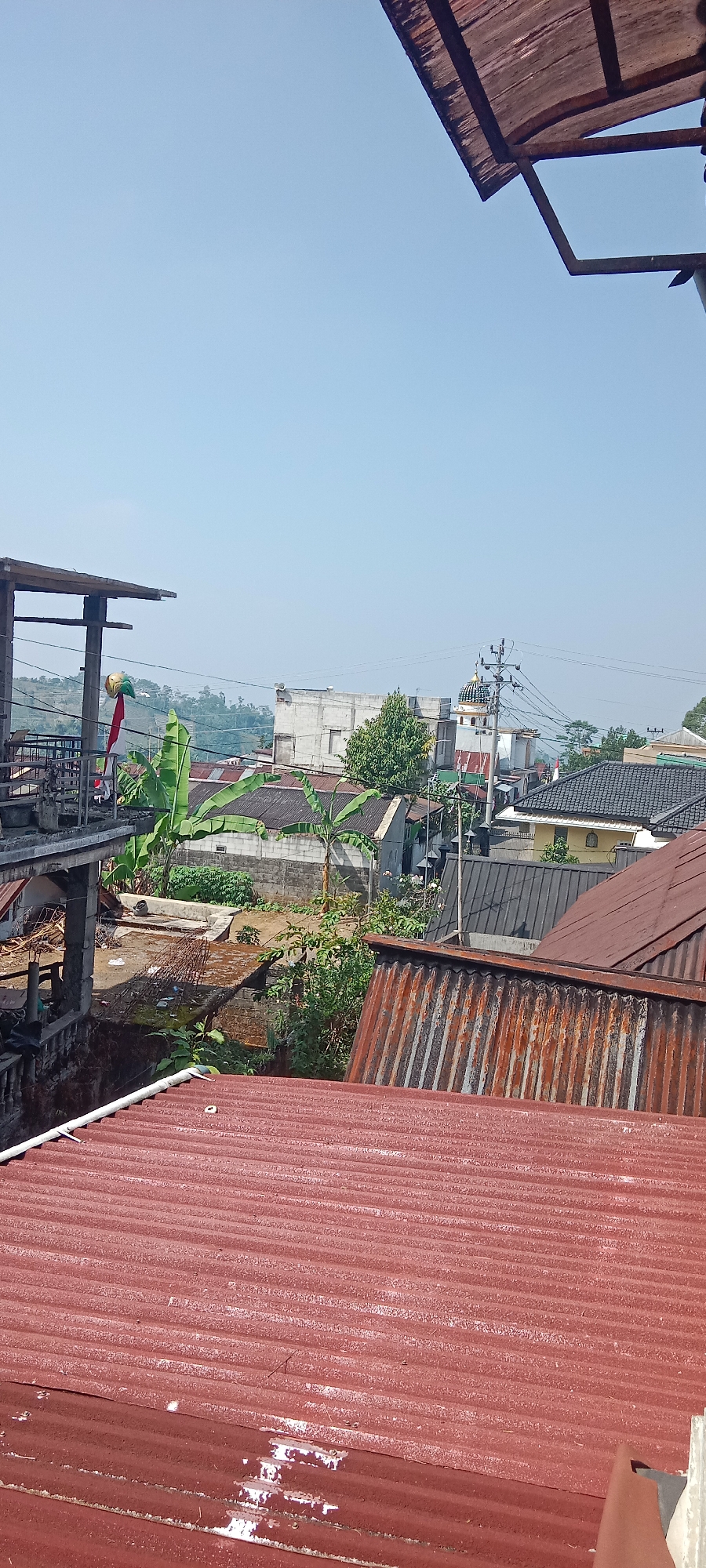 This image captures a view from an elevated position overlooking a residential area in Krinjing, Indonesia, under a clear blue sky. In the foreground, a weathered, red corrugated metal roof dominates the lower portion of the frame. Beyond this, a second, older corrugated metal roof, also showing signs of rust and age, is visible.
Further into the scene, a construction site or unfinished building is partially visible on the left, with concrete pillars and a wooden overhang. A bright yellow and green inflatable object and a red flag are attached to a pole at this location. Lush green banana plants with large leaves are scattered throughout the scene, indicating a tropical climate.
The middle ground reveals a collection of houses with various roofing materials, including corrugated metal and darker tiled roofs. Some houses appear to be constructed with concrete walls. A mosque with a distinctive dome and minaret is visible amongst the buildings, suggesting the presence of a Muslim community. Power lines crisscross the scene, supported by utility poles. In the distance, rolling green hills are faintly visible under the bright sky. The overall impression is of a densely populated, somewhat informal urban or semi-urban environment.