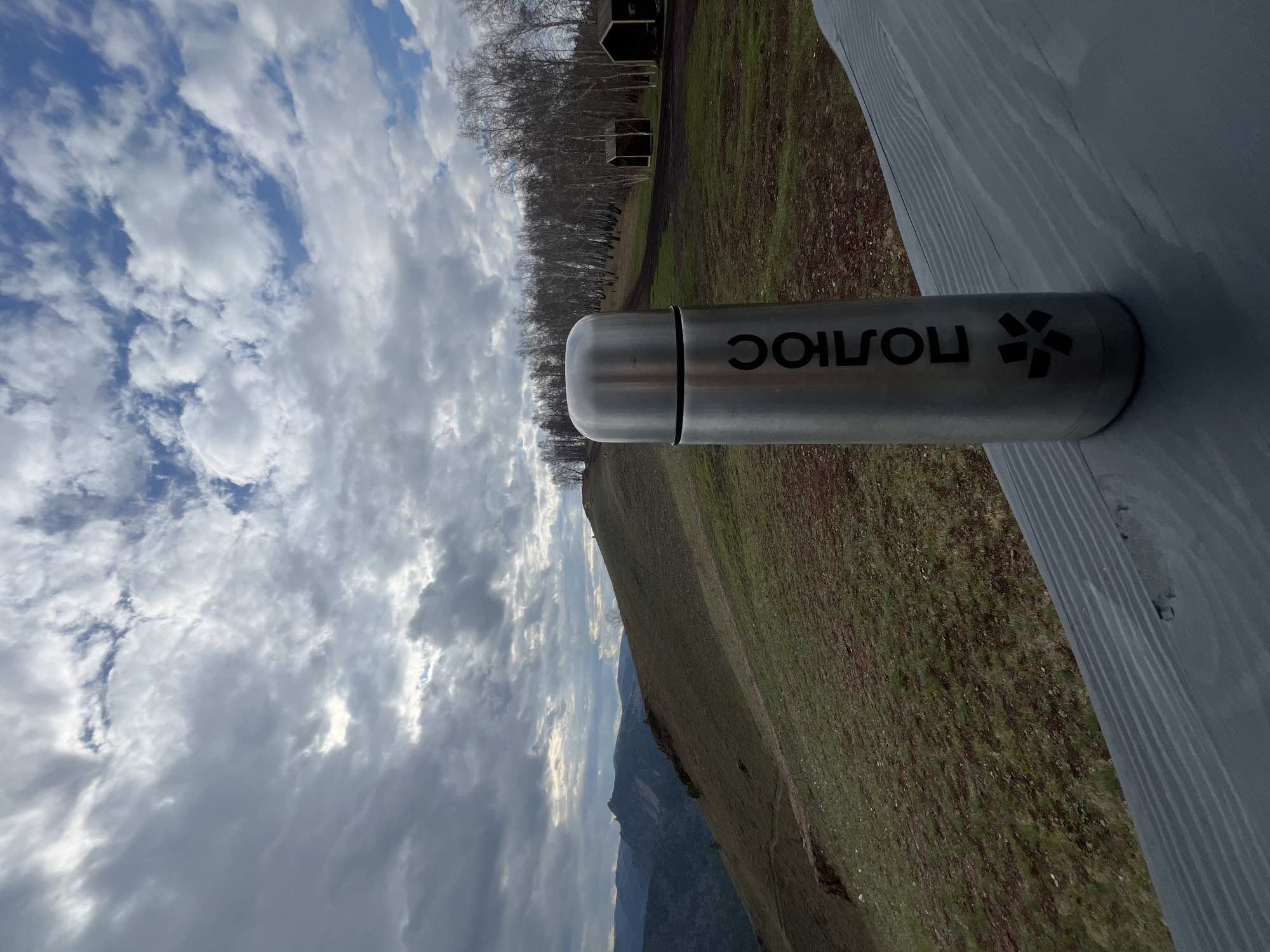 A silver thermos with black lettering sits on a gray wooden surface. The word "SOLOIUC" is printed backwards on the thermos. Behind the thermos, a grassy field slopes downwards toward mountains under a cloudy sky. In the distance, there are trees and small buildings. The location appears to be a park overlooking a mountainous landscape, possibly near Krasnoyarsk, Russia, based on the lettering on the thermos.