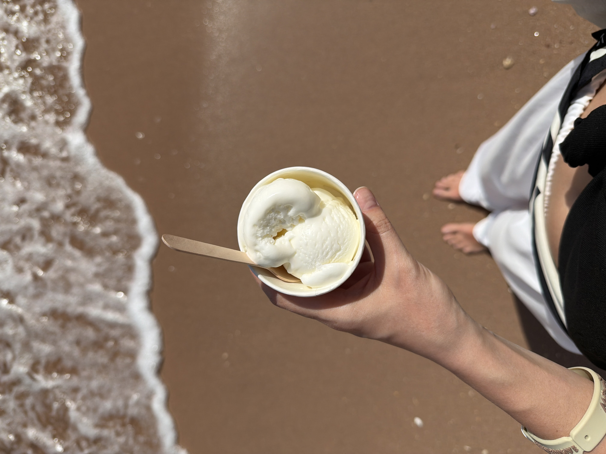 An overhead shot shows a person holding a cup of vanilla ice cream on a beach. The person is fair-skinned, wearing a patterned watch, white pants, and a black top. Their bare feet are visible on the sandy ground. In their hand, they hold a white paper cup filled with scoops of vanilla ice cream, accompanied by a wooden spoon. The beach setting includes light brown sand and the foamy edge of the ocean waves. The picture was taken in Sharm El Sheikh, Egypt.