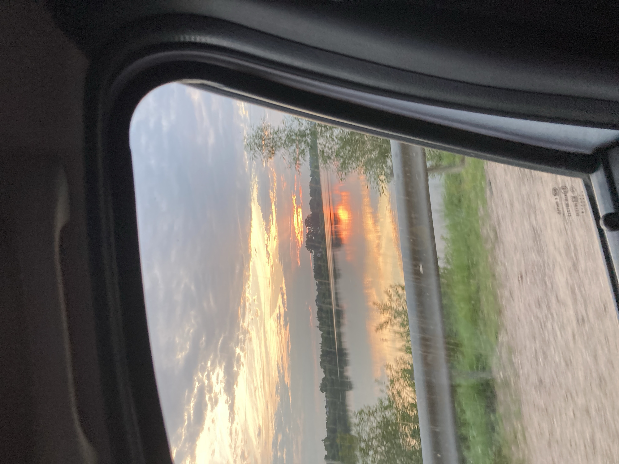 The image depicts a view from inside a vehicle, looking out through a car window. The scene outside is a landscape at sunset, with the sun low on the horizon, casting a warm glow across the sky and reflecting on a body of water. The sky is filled with dramatic clouds, tinged with orange, yellow, and pink hues. A line of trees is visible along the edge of the water. On the right side of the image, there is a dirt or gravel road and some green foliage, possibly grass or trees, partially obscured by a reflective barrier or part of the car's exterior. The interior of the car is visible in the foreground, with dark, textured surfaces framing the window.
The time of day is clearly sunset, indicated by the low sun and the colors in the sky. The weather appears to be clear with scattered clouds. The location is hinted at by a small piece of text on the car window, which appears to be a vehicle registration or compliance sticker, and has Cyrillic characters, consistent with the specified location of Niasvizh, Belarus. No people are visible in the image. The overall mood is peaceful and serene, capturing the beauty of a sunset over a natural landscape.