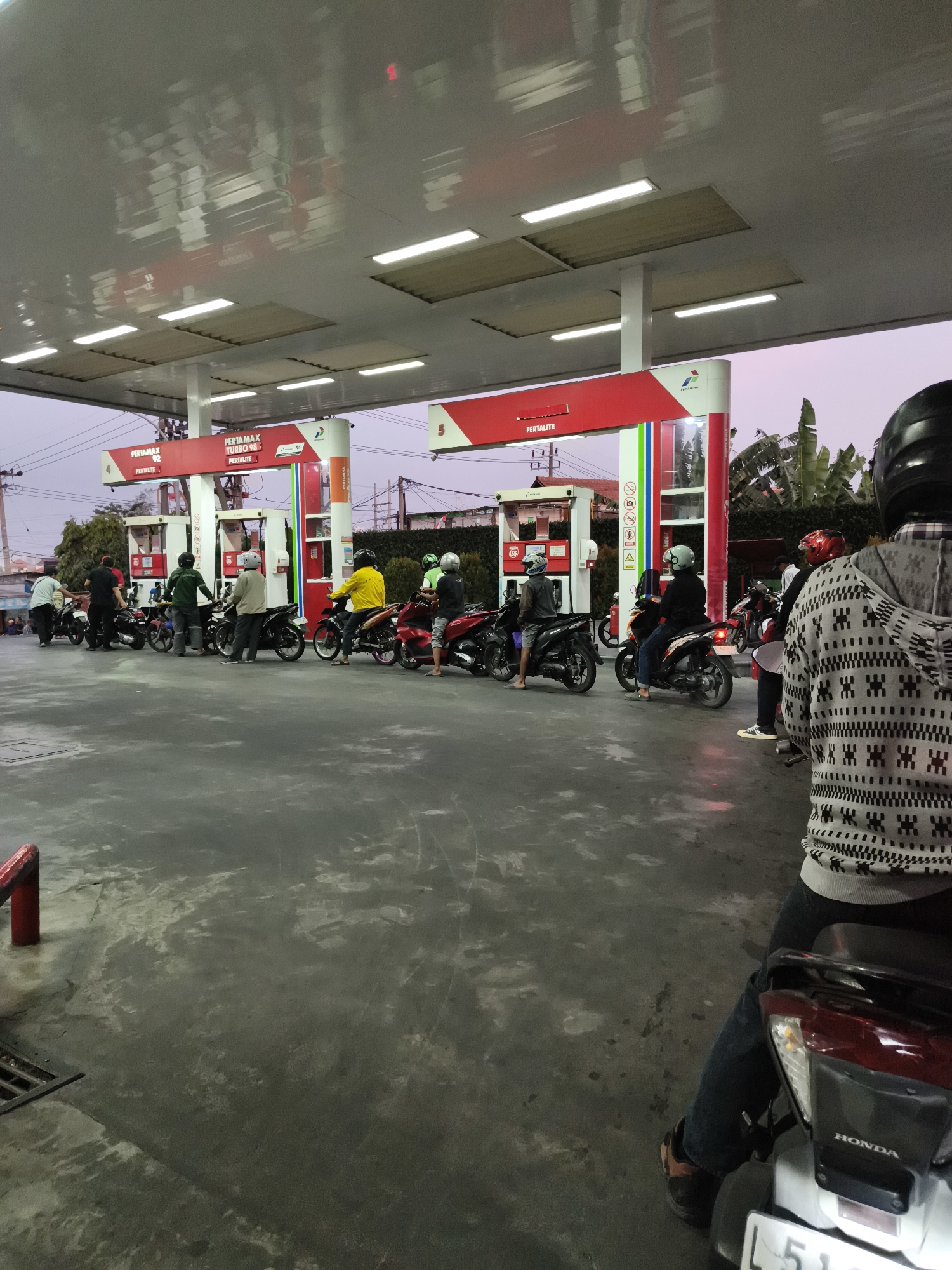 A wide shot captures an outdoor Pertamina gas station in Surabaya, Indonesia, at dusk. The sky is a pale purple. Multiple gas pumps, painted red and white with the Pertamina logo, are under a large, illuminated canopy. Several people on motorcycles are lined up at the pumps, either waiting or in the process of refueling. The ground is paved and appears damp. In the foreground, a person wearing a black helmet and a patterned grey sweater sits on a white Honda motorcycle. To the left, other individuals and motorcycles are visible, some obscured by the pumps. The overall scene depicts a busy, functional, everyday moment at a common urban service station.