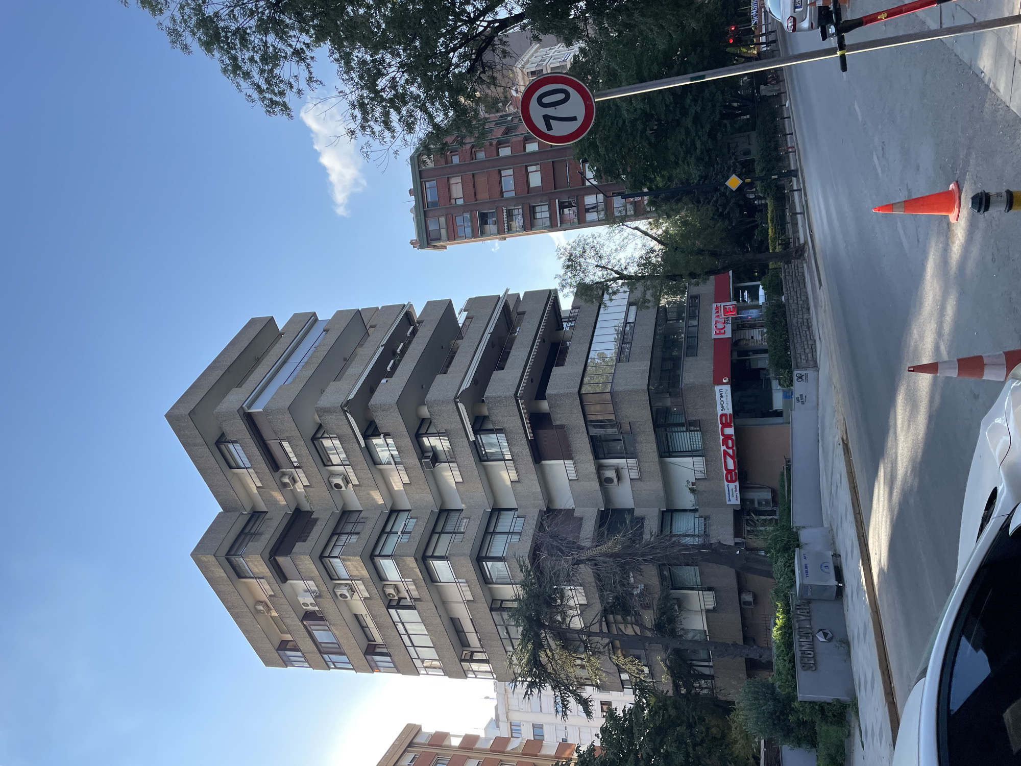The image depicts a street scene in Kadıköy, Turkey, on a clear, sunny day. A multi-story concrete apartment building with numerous balconies and windows dominates the left side of the frame. To the right of this building, a taller, brick-clad apartment building is visible. A red and white circular traffic sign indicating a speed limit of "70" is mounted on a pole in the foreground, to the right. Trees line the street and partially obscure the buildings. The street itself is paved, with a white car visible in the lower left corner and an electric scooter leaning against a pole on the right. In the foreground, several traffic cones are placed on the roadway. The ground level of the concrete building features commercial spaces, including a pharmacy with a prominent red sign reading "eczane".