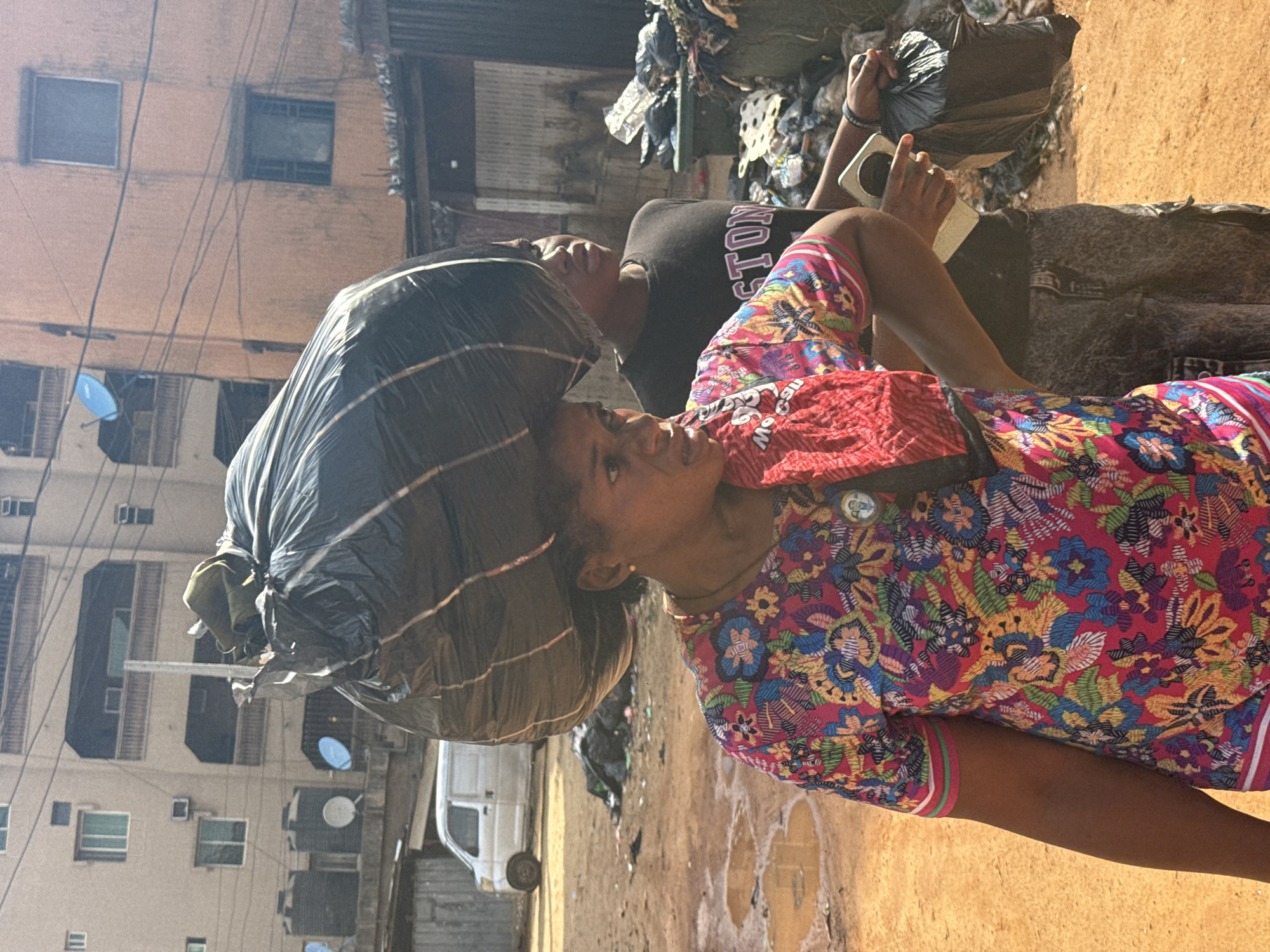 This image captures an outdoor street scene in Onitsha, Nigeria, under bright daylight. In the foreground, a woman carries a large, dark-colored plastic bag balanced on her head, a common method of transport. She is seen in profile, looking slightly upwards with a focused expression. She wears a vibrant, multi-colored short-sleeved dress adorned with intricate floral and abstract patterns in shades of pink, red, blue, green, and yellow. A red shoulder bag or pouch hangs across her chest.
Behind and to the left of the woman, a man stands, also looking upwards. He is wearing a black t-shirt with the word "BOSTON" visible in white lettering across the chest. He holds a light-colored mobile phone in his right hand.
The background reveals a densely packed urban environment. Multi-story buildings with various windows and what appear to be air conditioning units line the street. Numerous tangled black electrical or utility wires crisscross irregularly in front of the buildings. Small blue satellite dishes are visible on some building exteriors. The ground is unpaved, appearing as dirt or mud, with several puddles reflecting the bright sky. Piles of discarded items and debris are scattered on the ground and possibly on ledges of buildings, indicating a cluttered setting. A white van is partially visible parked in the distance. The scene suggests a busy, bustling atmosphere typical of a Nigerian city.