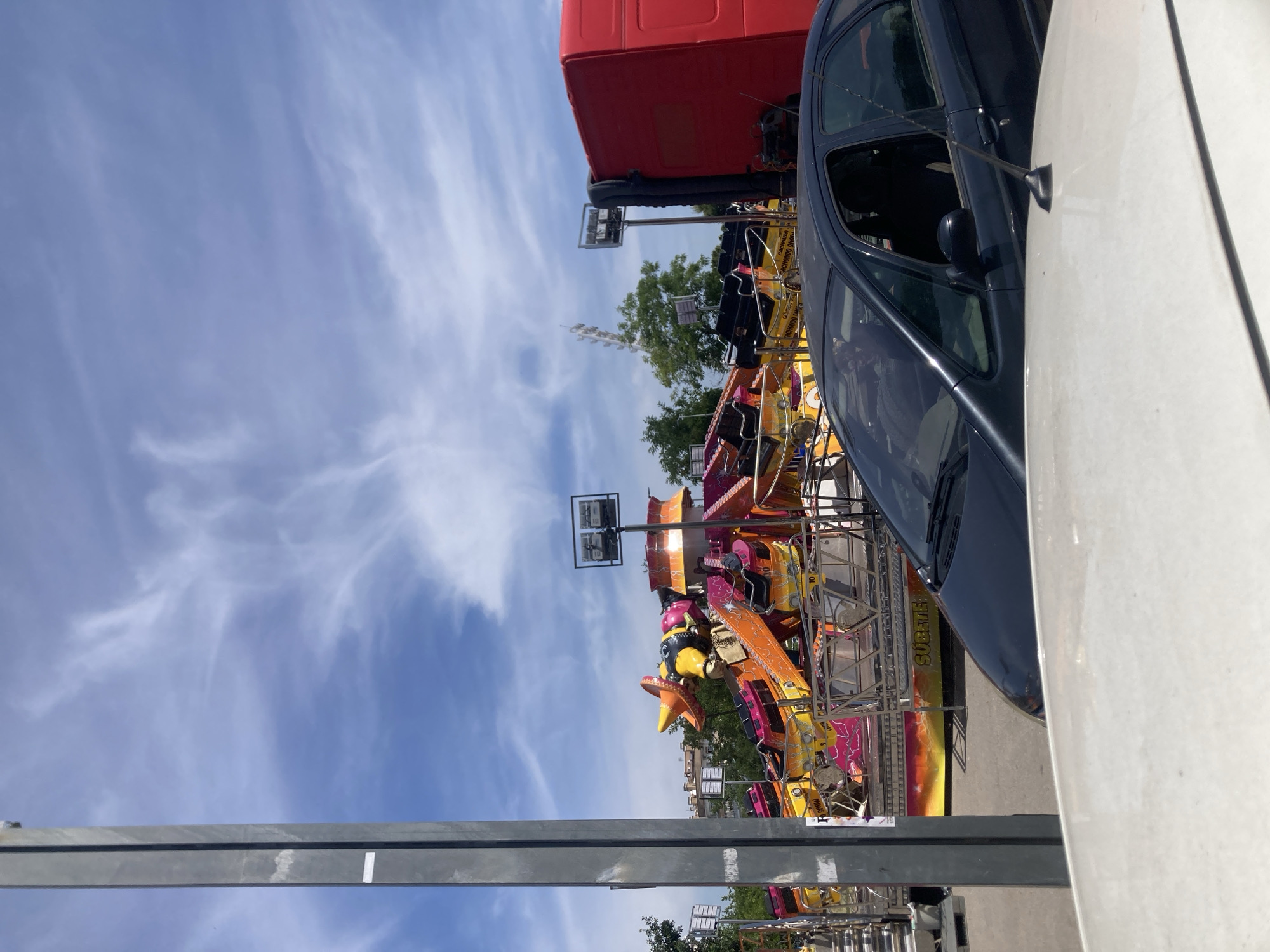 The image depicts an outdoor scene with a bright blue sky adorned with wispy white clouds. In the foreground, a tall, gray metal pole stands vertically on the left side of the frame. To the right of the pole, a colorful amusement park ride, predominantly in shades of pink, orange, and yellow, is visible. This ride features multiple seats and appears to be themed with a character wearing a sombrero. Behind the amusement ride, a red truck is partially visible on the right edge of the image. In the middle ground, a dark-colored car is parked, obstructing a clear view of the amusement ride's base. The ground appears to be paved. In the distance, trees and buildings are visible, suggesting an urban or suburban setting. The presence of the amusement ride strongly suggests a temporary fair or festival. This scene is located in the city of Terrassa, Spain.