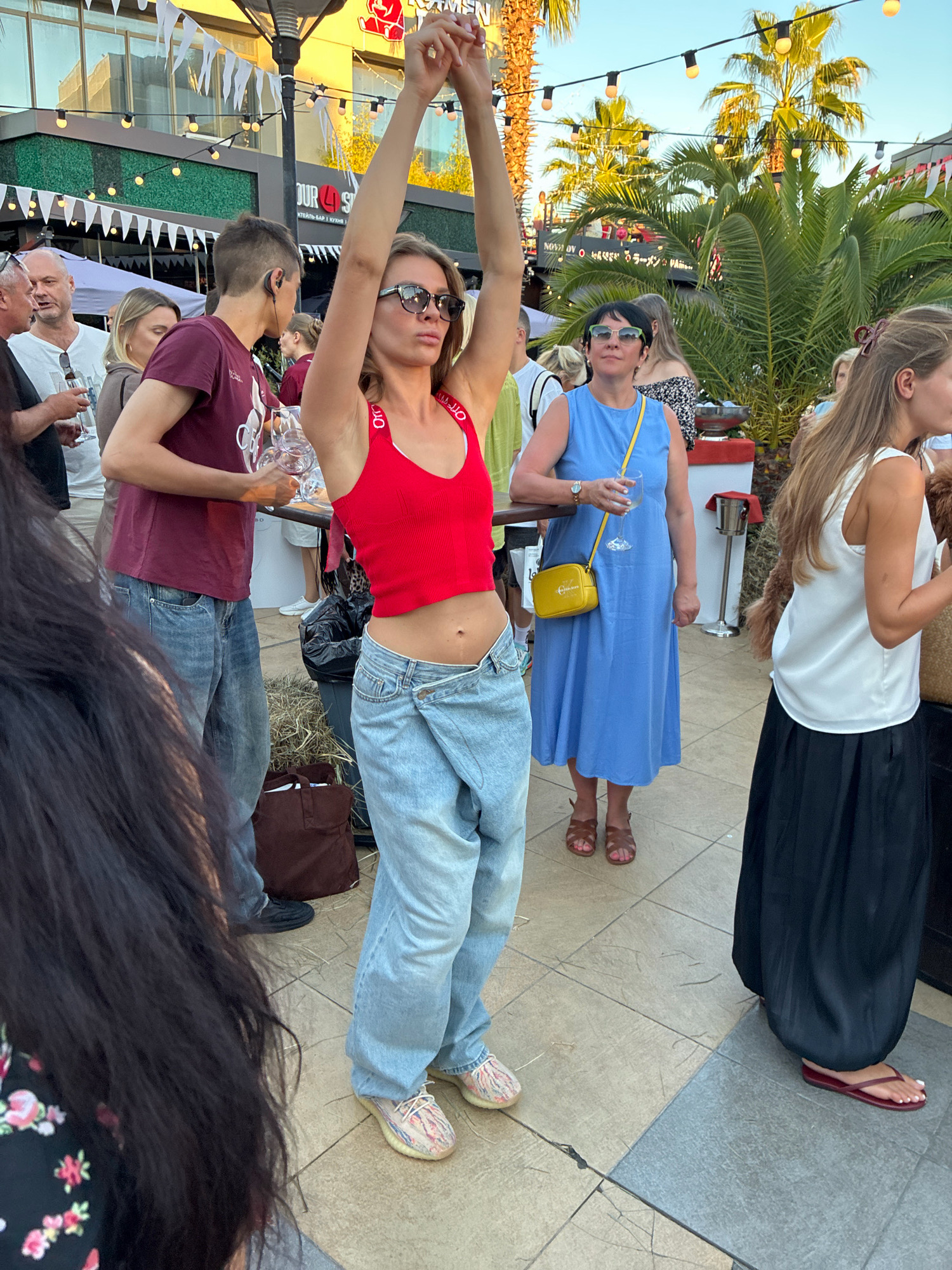 A medium-shot, eye-level, outdoor photograph captures a group of people at what appears to be a daytime outdoor event. In the foreground, a woman in a red tank top and baggy, light-wash jeans with a draped front is captured mid-motion, her arms raised above her head. She is wearing sunglasses and her navel is exposed. To her left, a man in a burgundy t-shirt and jeans holds a few empty wine glasses. Behind them, other individuals are visible, including a man in a white t-shirt and a woman in a blue dress holding a glass of white wine and a yellow crossbody bag. In the background, palm trees, string lights, and building facades suggest a warm-weather, possibly resort-like setting. The ground is paved with light-colored tiles. The scene is likely from the city of Sochi, Russia, given the architecture and vegetation often found in its resort areas.