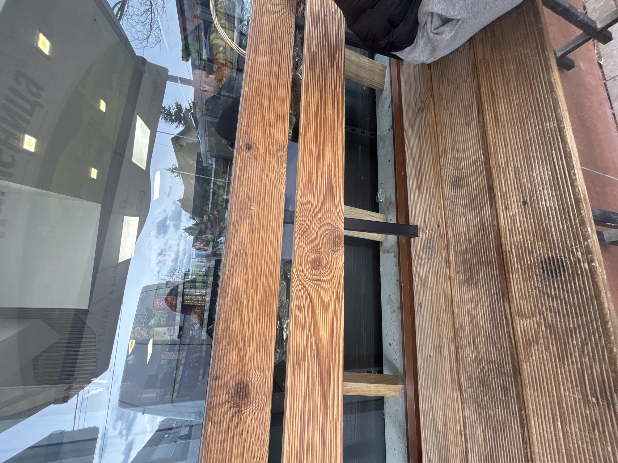 The image shows a wooden bench located outdoors, next to a shop window. The shop window has lettering on it. Outside, there is a vending machine. The overall scene appears to be within an urban environment. Without additional information, pinpointing the precise location in Plovdiv is impossible. The perspective is shot from above. There is a person in the upper right of the frame.