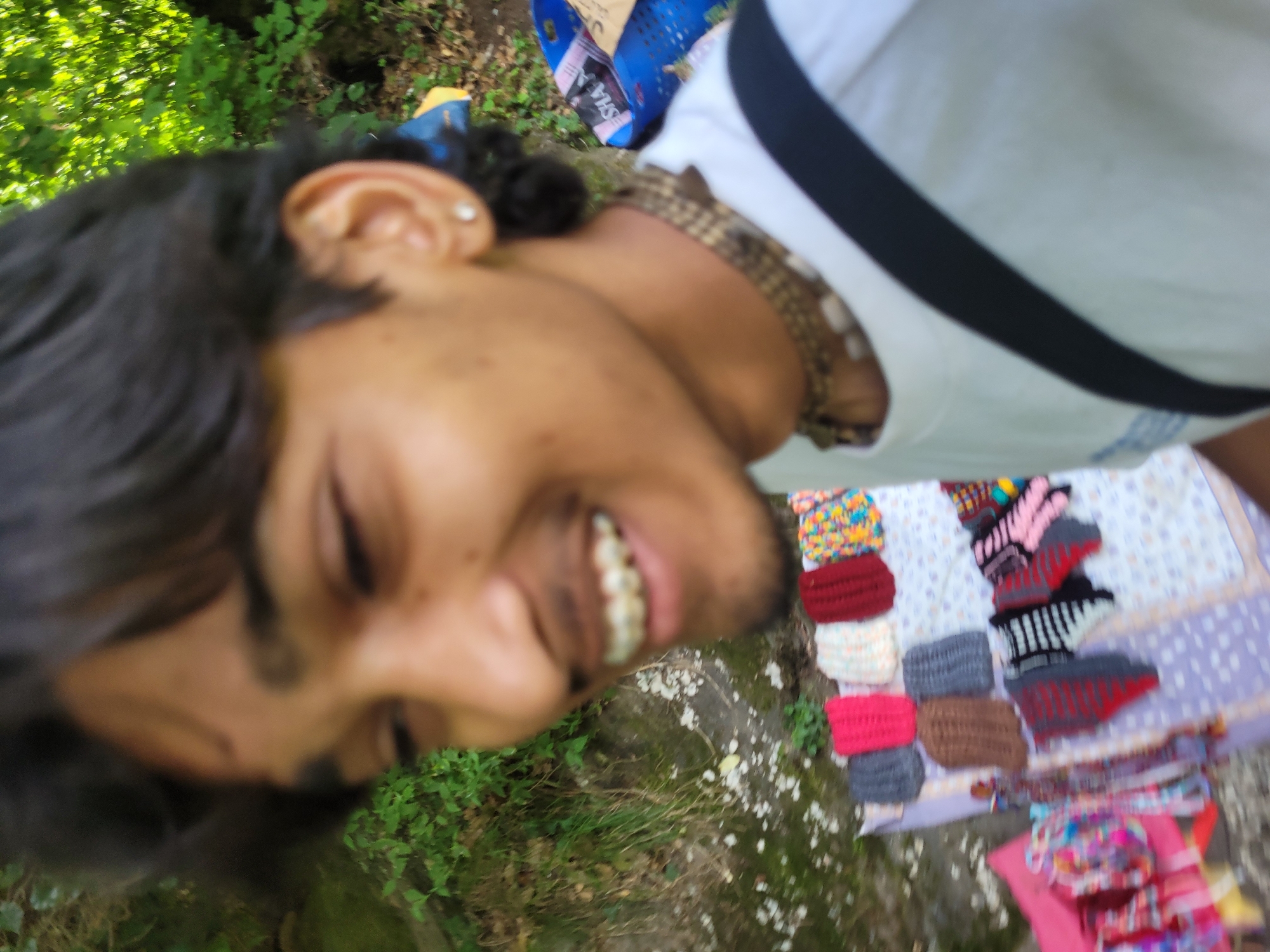 The image shows a close-up of a young man with dark hair, smiling and looking to the left. He is wearing a white t-shirt and a beaded necklace. A black strap crosses his chest. In the background, to the left, is lush green foliage. Below him, a table is covered with a patterned cloth, displaying various handmade knitted items, including hats and other small accessories in vibrant colors like pink, red, brown, grey, and multicolored patterns. Some of these items are arranged in rows, while others are displayed on a pink cloth to the left. A blue basket with some items and the word "SHA" visible is also present in the background, suggesting a market or street stall setting. The overall impression is that of a vibrant outdoor scene, possibly in a place like Vashisht, India, known for its handicrafts and natural beauty.
