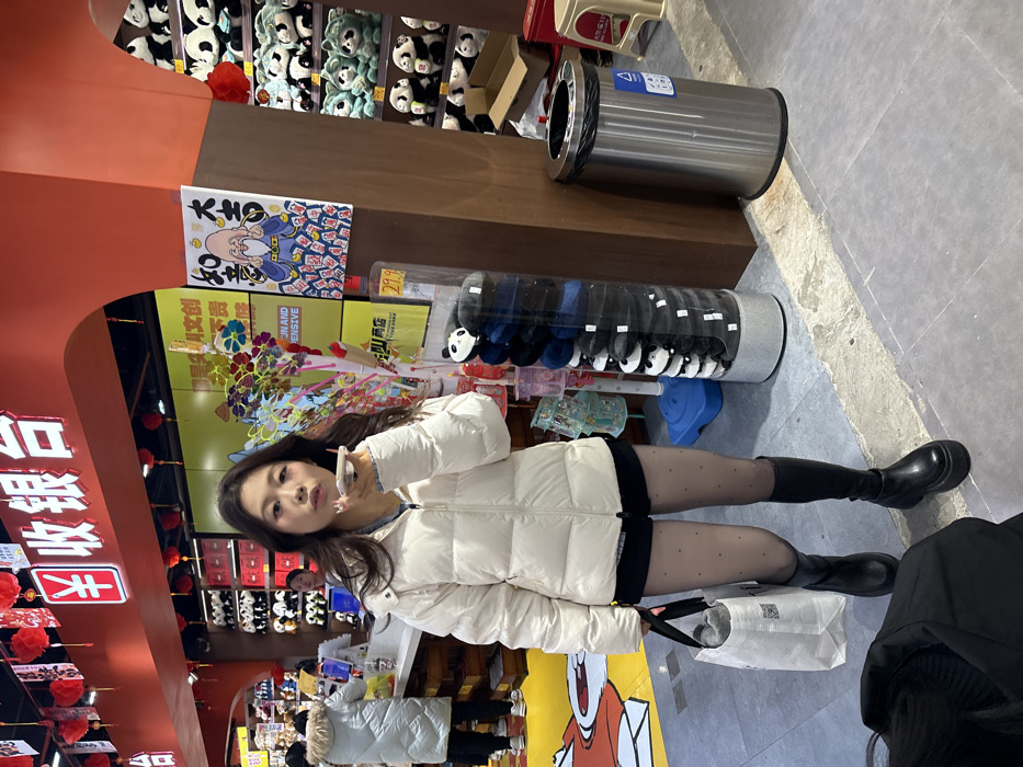 The scene depicts an indoor retail environment located in Yuzhong District, China. A female individual with long dark hair stands centrally, facing forward, holding a light-colored smartphone to her mouth, consistent with speaking or recording audio. She is attired in a cream-colored puffer jacket, black shorts, sheer black tights with small patterned dots, and black platform knee-high boots. She carries a white fabric tote bag with black handles and a visible QR code in her left hand.
The background features a bright retail space with a red overhead structure displaying white Chinese characters "¥ 收银台" (Cashier). Multiple red lanterns hang from the ceiling. To the right of the main subject, a tall, clear cylindrical display stand contains a stack of black berets or hats, each featuring a white panda face. Adjacent to this, a large poster depicts a stylized elderly figure surrounded by playing cards or tiles, with Chinese characters "大吉 如意" (Good fortune, all wishes fulfilled) and "发" (wealth/prosperity). Another colorful poster nearby is partially visible, showing English text including "FUN AND EXPENSIVE". A stainless steel cylindrical trash can with a black lid is positioned to the far right.
In the left background, a light-colored counter is present where two other individuals are partially visible; one in a white puffer jacket and another appearing to be a store employee. Shelves lining the walls behind the counter are stocked with numerous plush toys, primarily panda bears and some green animal figures. The floor consists of light gray tiles with sections painted yellow and red, including cartoon-like illustrations.