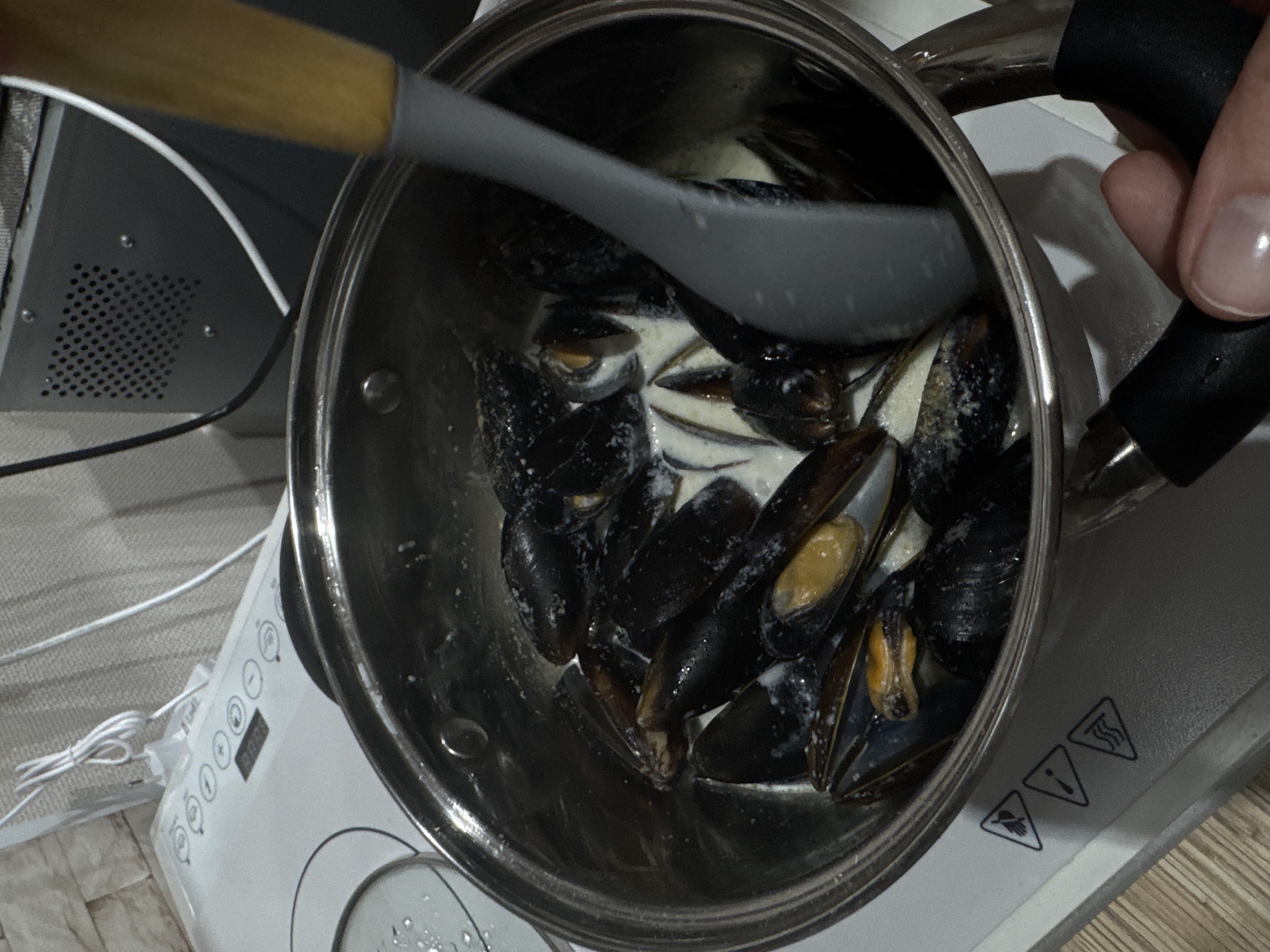 A top-down shot depicts a stainless steel pot filled with cooked mussels in a creamy white sauce. A gray cooking utensil with a wooden handle is stirring the mussels. A hand is holding the pot by one of its black handles. The pot is sitting on a white induction cooktop. In the background, a gray appliance is partially visible. The scene is likely indoors in a kitchen setting, although the specific location in Perm, Russia, cannot be determined from the image.