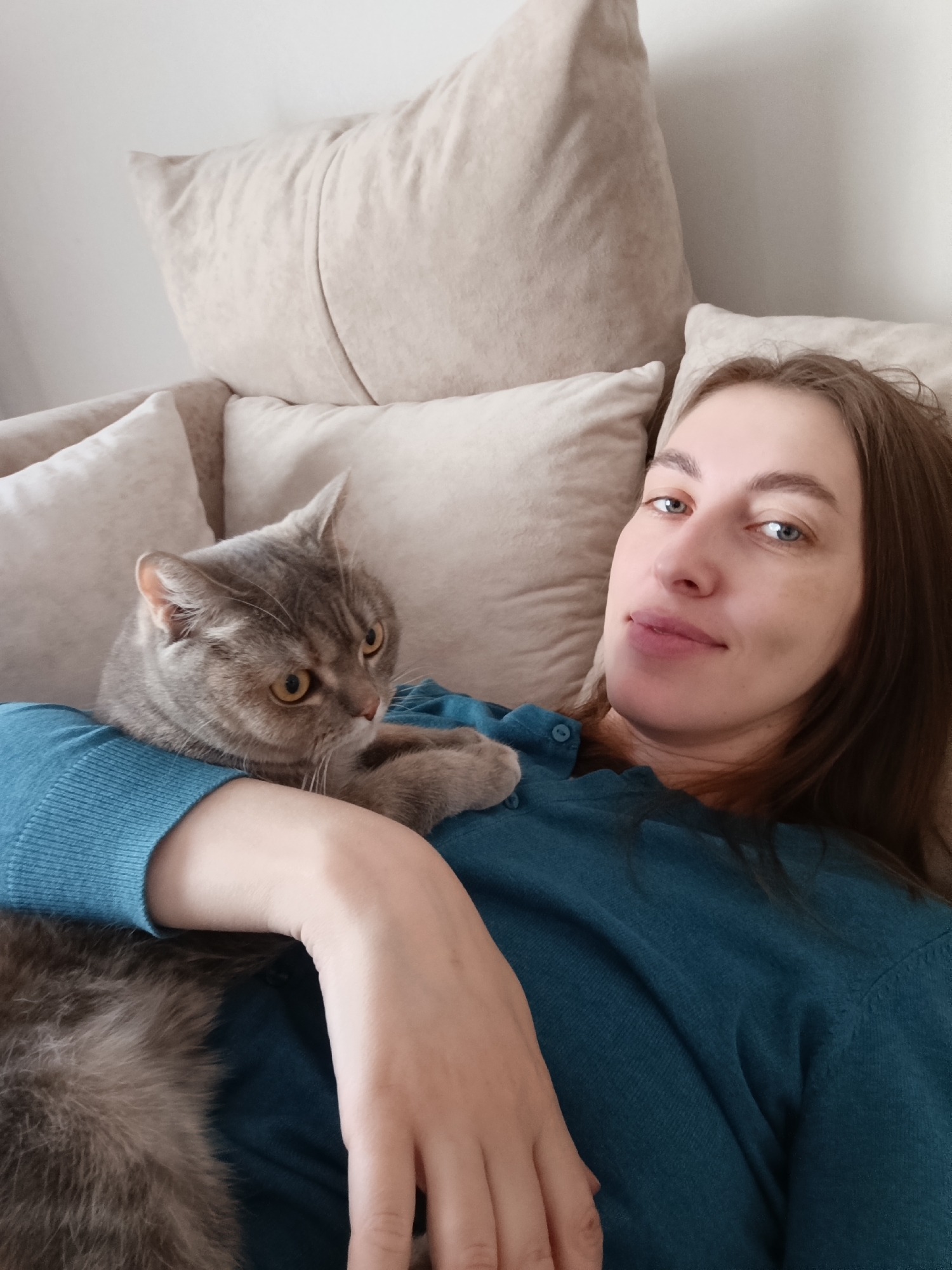 This indoor image features a woman and her cat sharing a quiet moment, likely within a home in Chaikovsky, Russia. A young to middle-aged woman with long brown hair and blue eyes is reclining on a sofa or bed, supported by several plush, light beige pillows. She wears a long-sleeved teal or turquoise top and gazes calmly at the camera with a subtle, content smile.
Resting comfortably on her chest and left arm is a grey tabby cat, possibly a British Shorthair, with prominent stripes and bright amber eyes. The cat is looking off to the viewer's left, appearing alert yet relaxed, with one paw gently placed on the woman's arm. The lighting is soft and natural, indicating daytime. The scene conveys a sense of peaceful companionship and domestic comfort. There are no visible texts or specific location cues beyond the general indoor setting.