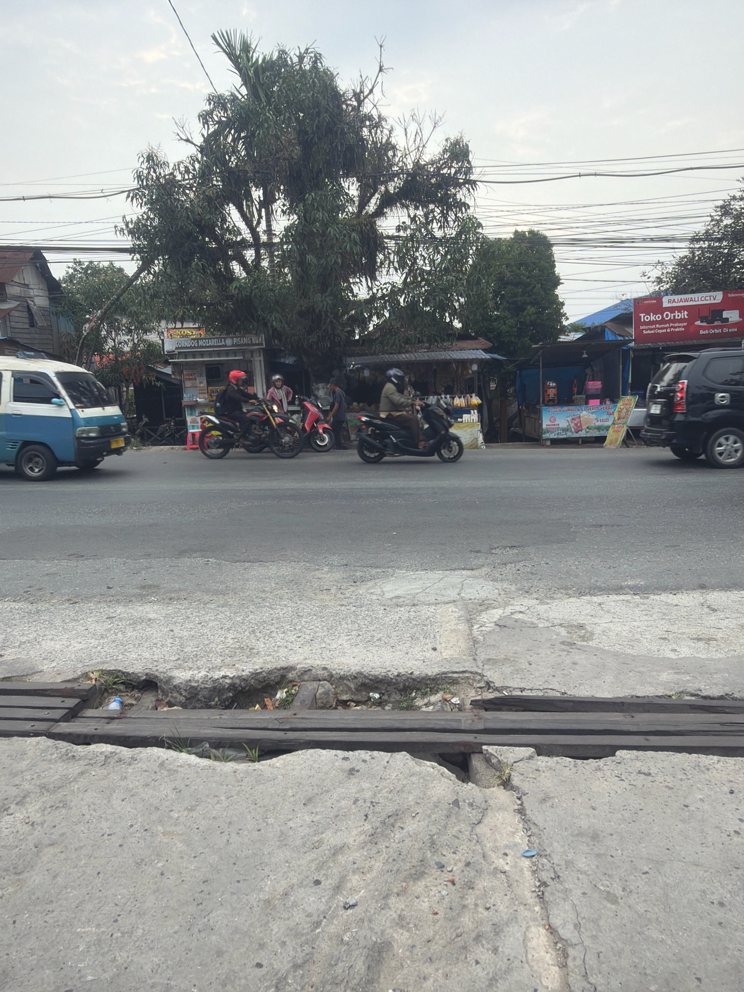 The image captures a street scene in Balikpapan, Indonesia, under an overcast sky. A paved road occupies the foreground, with a damaged concrete drainage culvert running across it, reinforced by wooden planks. Beyond the road, several small businesses and stalls line the street, some with awnings providing shade.
On the road, a light blue and white van is traveling from left to right. Several individuals on motorcycles are also present. One motorcycle, ridden by a person wearing a red helmet and a black jacket, is moving from left to right, passing a pink motorcycle. Another person on a motorcycle, wearing a helmet and a light-colored jacket, is moving from right to left. A black SUV is parked on the right side of the road, with its rear visible.
One of the businesses has a prominent sign that reads "Toko Orbit" and "RAJAWALICCTV". Another stall displays signs related to food items like "CORN DOG MOZZARELLA" and "PISANG KUY". The overall environment suggests a commercial area with active street traffic and roadside vendors. A large tree with dense foliage stands behind some of the stalls, and power lines are visible crisscrossing the sky.