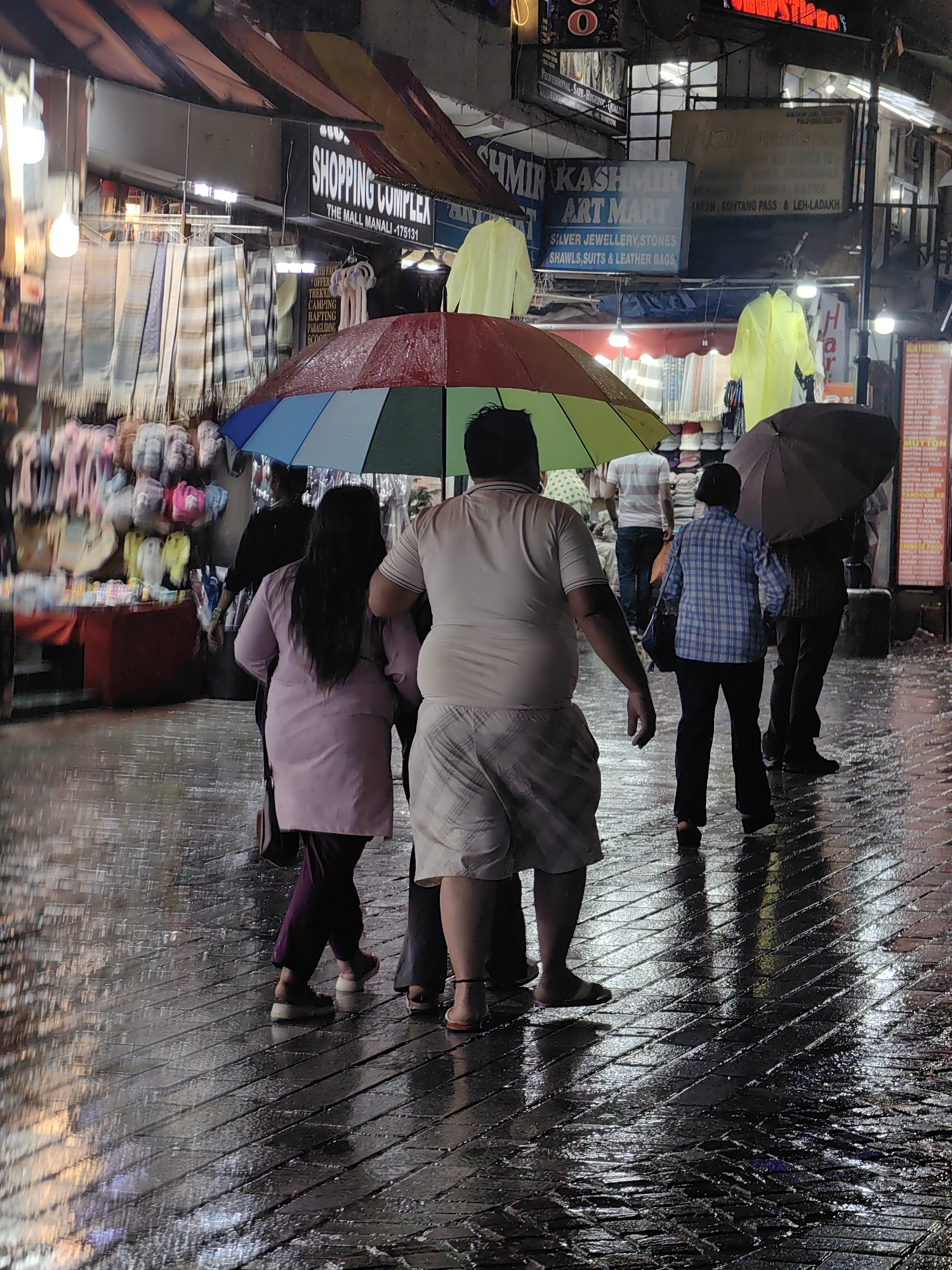 The image depicts a rainy evening scene at the Mall Road in Manali, India. Several people are walking on the wet, reflective cobblestone street, under umbrellas. In the background, shops line the street, with various goods displayed, including textiles and clothing. Signs for "Shopping Complex," "Kashmir Art Mart," and restaurant menus are visible. The atmosphere is illuminated by artificial lights, casting reflections on the wet ground. The people are dressed in casual attire, and some are carrying bags. One person in the foreground is wearing a light-colored t-shirt and checkered shorts. Another person in the middle is wearing a pink jacket and dark pants. A person in a plaid shirt is also visible. The overall impression is that of a bustling marketplace during a downpour.