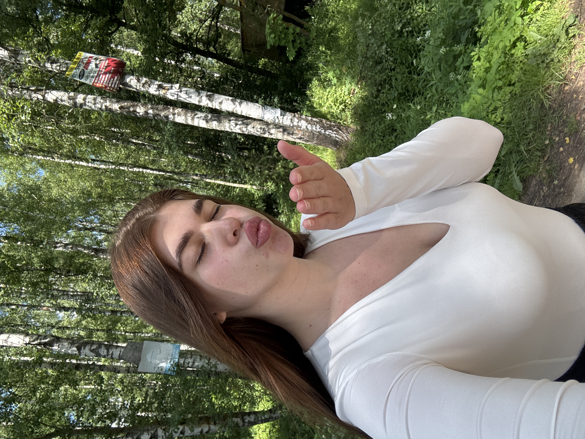 A young woman with long, brown hair and closed eyes is blowing a kiss towards the camera. She is wearing a white, long-sleeved shirt with a deep V-neck. Her hand is held up near her face, fingers extended, as if to emphasize the gesture. The background consists of a sun-dappled forest with tall birch trees. On one of the trees to the right, a red and white sign is attached. The ground is covered with green grass and some dry leaves. The location is identified as the city of Mechyory, Russia.