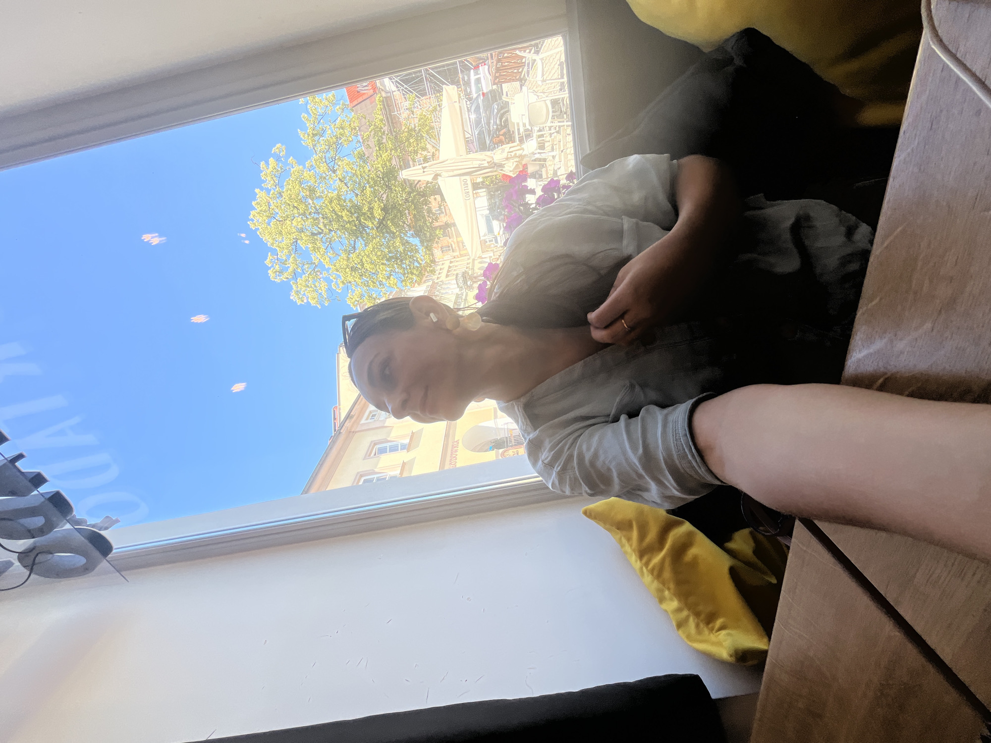 This media file is a horizontal, eye-level, indoor shot of a person looking out of a window. The person, whose face is partially obscured, is wearing a light-colored shirt and is positioned on the right side of the frame, leaning their head on their arm. They appear relaxed, with their gaze directed outward.
The setting is an indoor space with a large window that offers a view of an outdoor scene. The window has a white frame and seems to be situated at a slight upward angle, leading the viewer's eye towards the sky. Outside, the sky is a clear, bright blue, suggesting a sunny day. There are several blurry, light-colored objects floating in the sky, possibly bokeh effects or small, distant objects. A tree with vibrant green foliage is visible on the left side of the outdoor view, and beyond that, buildings with architectural details can be seen, including some with red tile roofs and windows. Some scaffolding or construction equipment is also present in the background.
The interior space is dimly lit, with the natural light from the window illuminating the scene. On the right side of the frame, part of a wooden surface, possibly a table, is visible, along with a brown or dark-colored cushion. To the left of the person, a bright yellow cushion is visible, and further left, some grey, rounded objects with wires, possibly electronic devices or accessories, are attached to the window frame.
The overall mood appears to be one of quiet observation or contemplation. The bright sunlight outside contrasts with the softer interior lighting. There is no visible text in the image. The time of day is likely daytime, given the bright, clear sky. The weather appears to be pleasant and sunny.