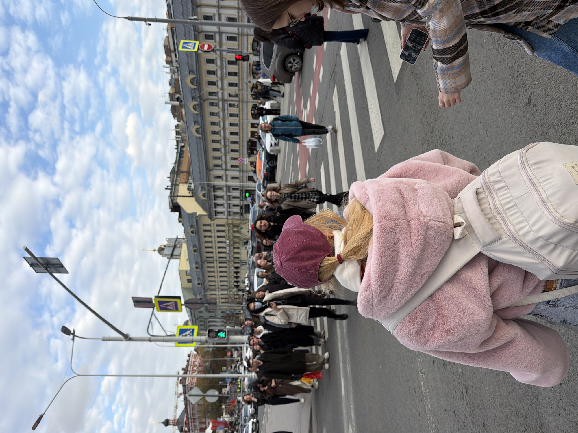 The image depicts a bustling urban street scene in Moscow, Russia, taken from an elevated perspective looking down a street with a pedestrian crossing. In the immediate foreground, the back of a person is visible, wearing a fluffy pink hooded jacket, a maroon baseball cap, and carrying a light-colored backpack. Their blonde hair is tied back in a ponytail.
To the right, another individual is partially seen, dressed in a plaid coat and blue jeans, holding a smartphone. They, along with numerous other pedestrians, are situated on or near a zebra-striped crosswalk in the midground, suggesting an active crossing or waiting period. Several cars are also visible on the street.
The background features a row of multi-story, classical-style buildings lining the street under a partly cloudy sky. Street furniture, including lamp posts, traffic lights (one showing red, another green), and various road signs, are prominent. Notable signs include a yellow square with a blue pedestrian crossing symbol and a red circular "no entry" or "stop" sign.
The scene captures a typical daytime moment in a city, with people moving about. Faint, partially obscured text, possibly "HOME YOUT...", is visible on the foreground backpack.