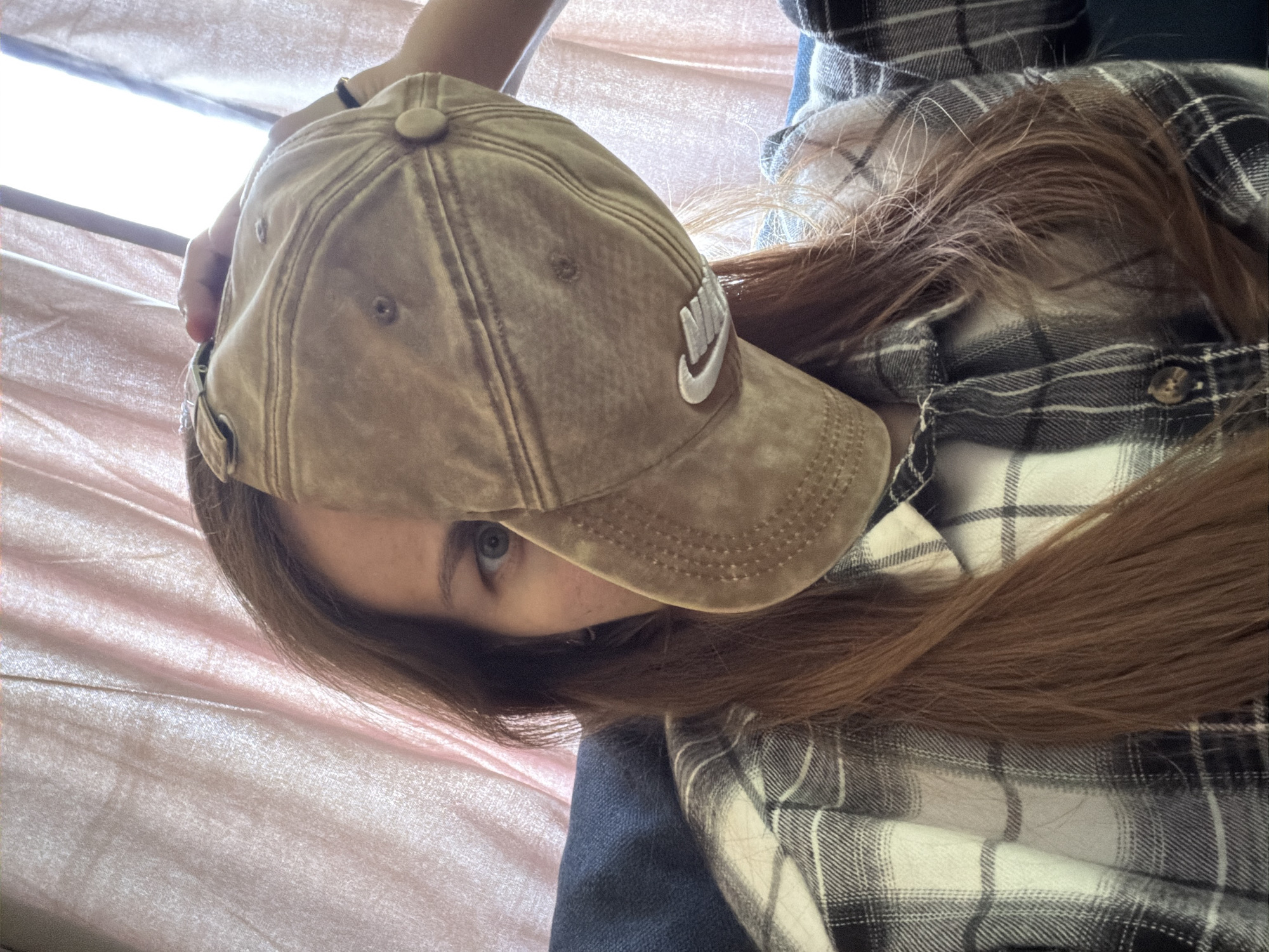 A young woman with long, brown hair is sitting on a couch and holding a tan baseball cap in front of her face, obscuring most of it. Her left eye is visible, and she appears to be looking directly at the camera. She is wearing a black and white plaid shirt. The background consists of a light pink, textured curtain and a dark blue couch. The image does not provide any information about the location in the city of Tambov, Russia.