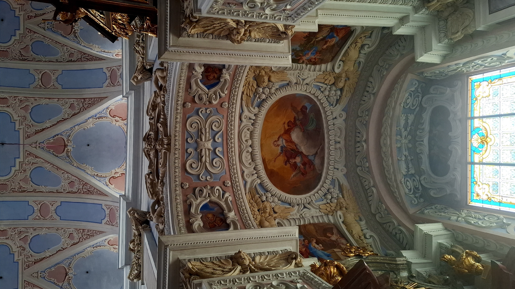 This is a low-angle, eye-level shot of a highly ornate, gilded and painted ceiling inside a church. The ceiling features elaborate frescoes and stucco work in pastel colors of pink, blue, and white, with gold accents. In the center of the image, an oval fresco depicts a woman in orange robes, surrounded by cherubs and angelic figures, ascending into a bright, radiant sky. Surrounding this central fresco are intricate stucco moldings with carved figures of angels and other decorative elements. Below this, an archway reveals more painted details and a stained-glass window with a vibrant blue and yellow design. The architectural style suggests a Baroque or Rococo influence. The image was taken inside the church of Maria Himmelfahrt in Furth bei Göttweig, Austria.