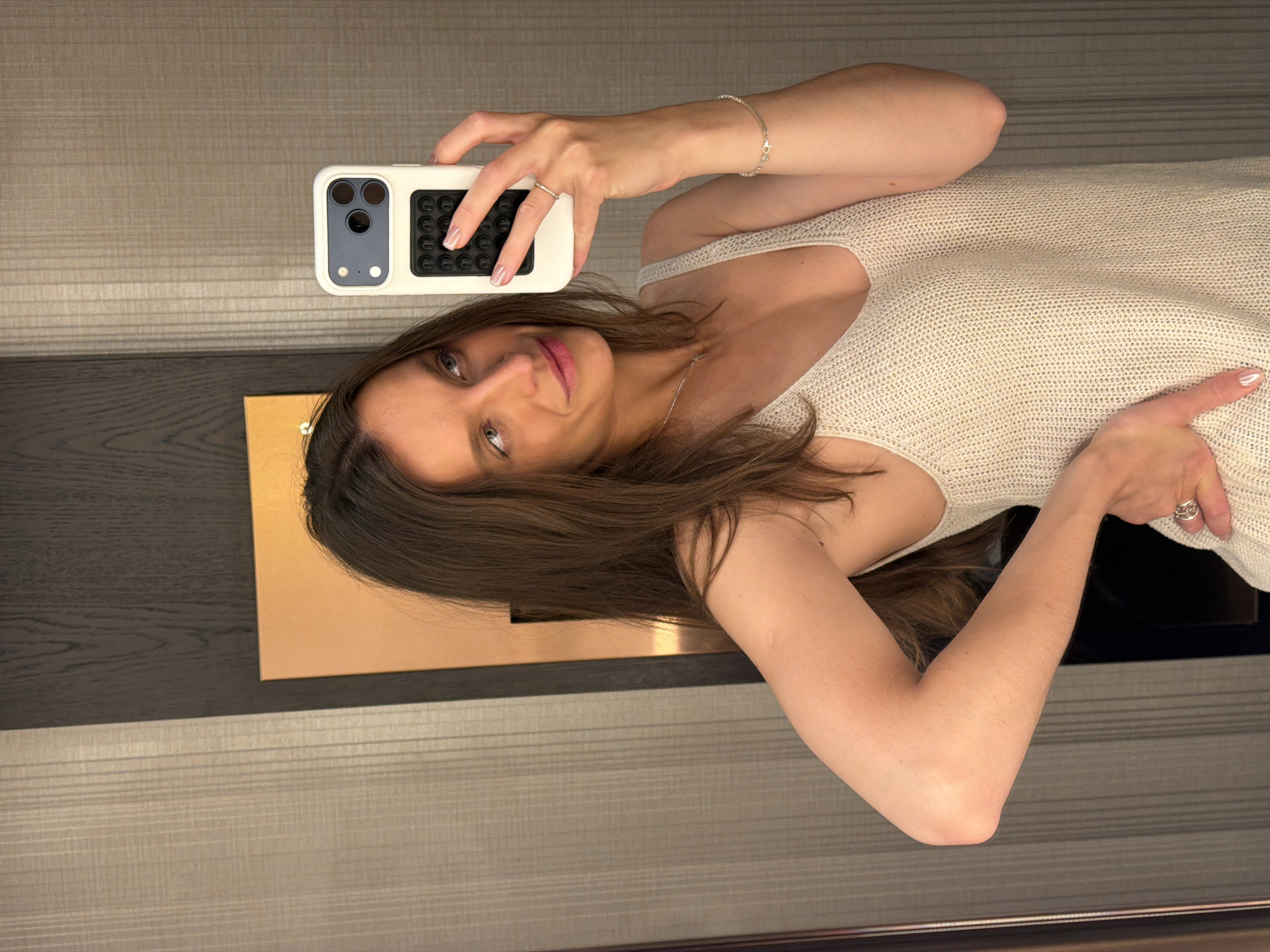 A female subject is captured in a mirror selfie, positioned indoors in what appears to be a hotel room or similar private space in Budva, Montenegro. The subject, with long, dark brown hair and light-colored eyes, is seen from the chest up, slightly angled to her right while looking up and to her left, displaying a subtle smile. She wears a light-colored, knitted V-neck tank top.
Her right hand holds a white smartphone, consistent with an iPhone Pro Max model, encased in a white protective cover featuring a black "pop it" style grip accessory on its back. A thin, silver-colored band ring is visible on the ring finger of her right hand. Her left arm is bent at the elbow, with her hand resting on her hip, revealing a wider, silver-colored ring on her left ring finger. A delicate, silver-colored tennis bracelet adorns her left wrist, and a thin silver-colored necklace with a small pendant is worn around her neck. Her fingernails are manicured with a light, possibly pearlescent polish.
The background, reflected in the mirror, shows a wall with vertical stripes or textured paneling in a light beige tone on the left side, transitioning to a darker, possibly wood-paneled or textured surface on the right. A rectangular, orange or bronze-colored panel is partially visible behind the subject's head on the right side. The lighting appears to be artificial.
