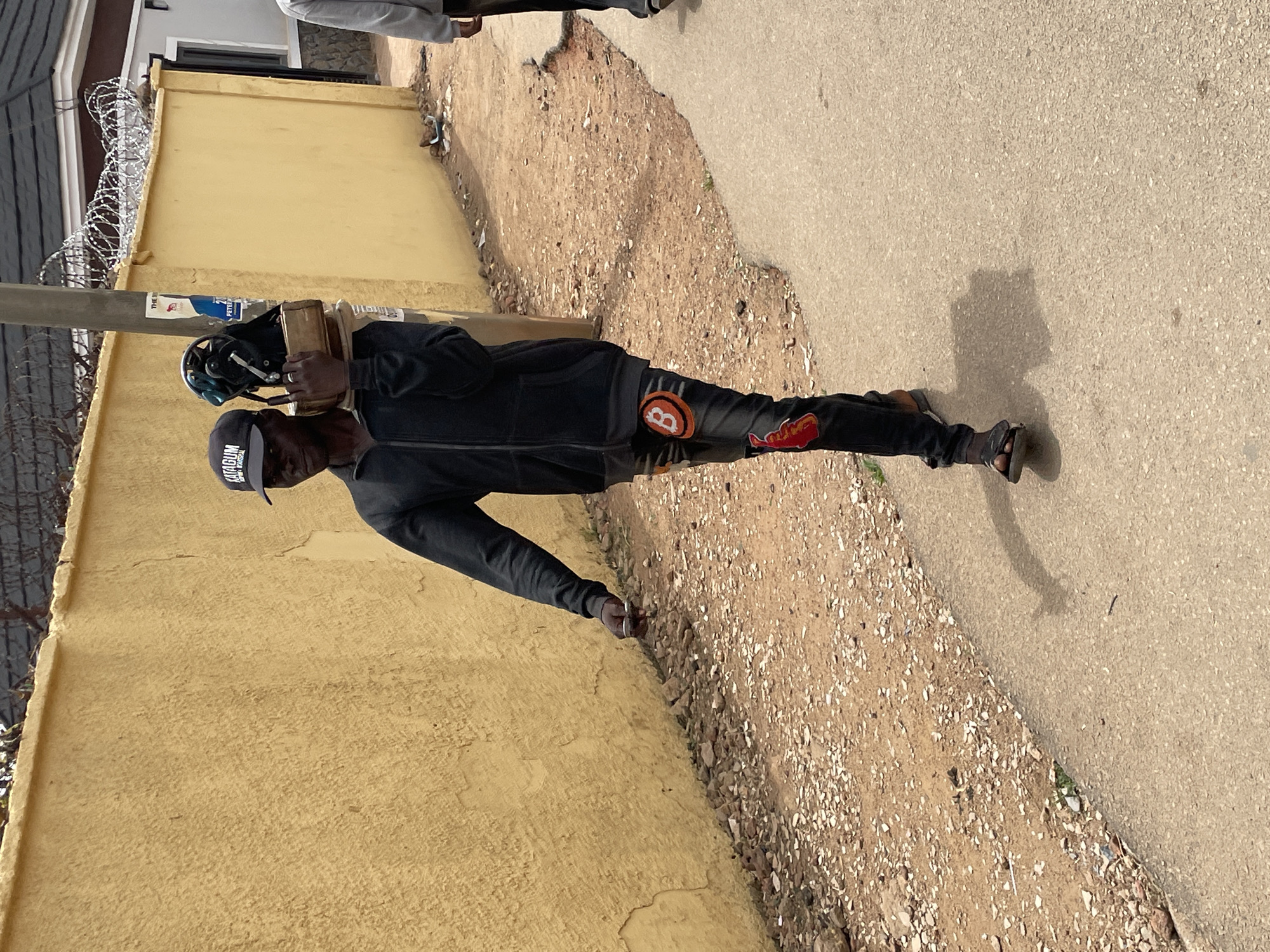 A man wearing a black hoodie, black pants with Bitcoin logos, and a black cap is walking down a street in Jos, Nigeria. He carries equipment on his shoulder. He is walking alongside a yellow wall topped with barbed wire. The environment is sunny. The ground consists of a combination of gravel and paved concrete.