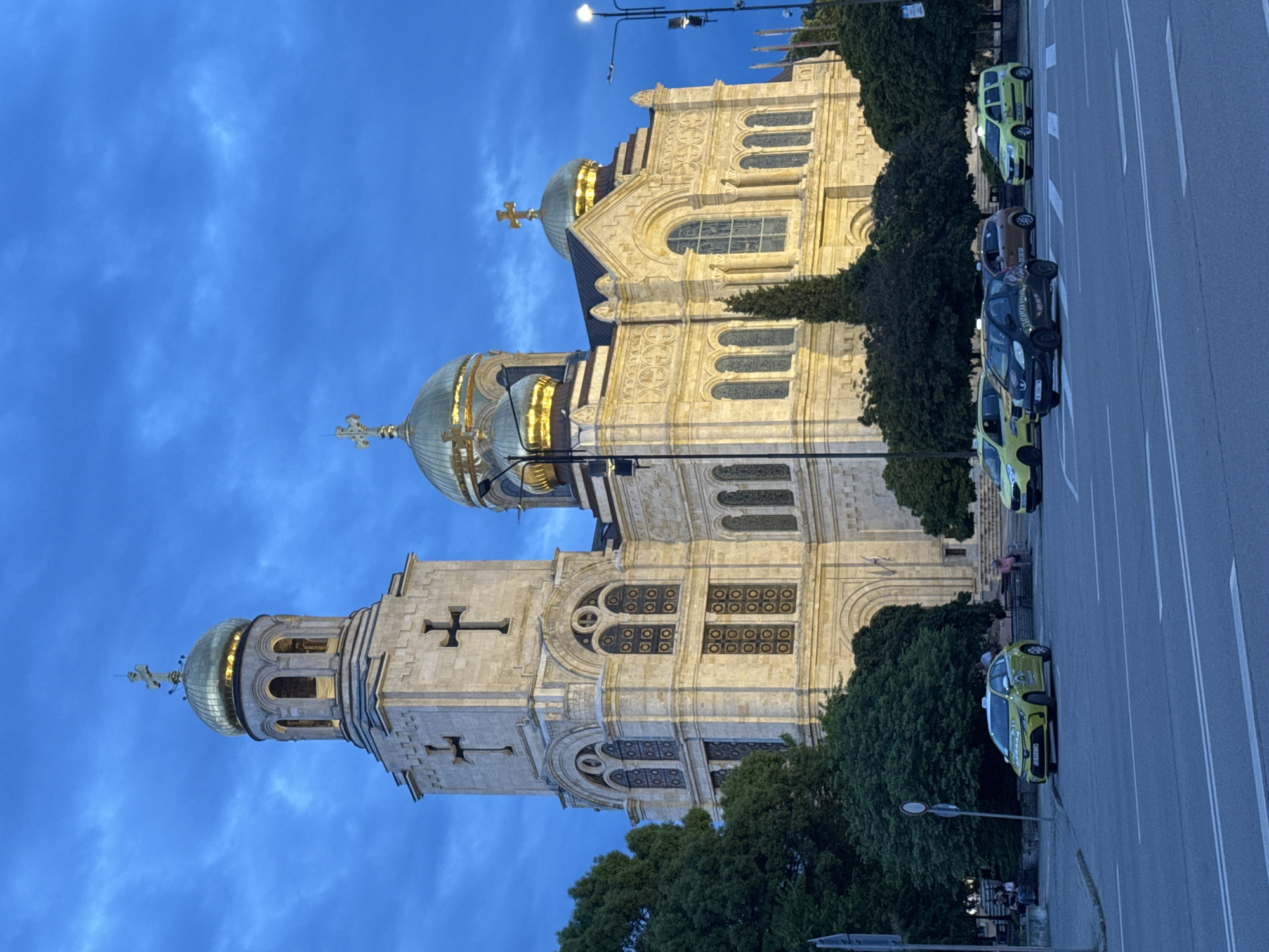 The image shows the Cathedral of the Assumption of the Virgin in Varna, Bulgaria, at dusk. The cathedral, illuminated with a golden hue, stands beside a road with several parked taxi vehicles. Trees line the base of the cathedral. The sky is a dark blue with some cloud cover.