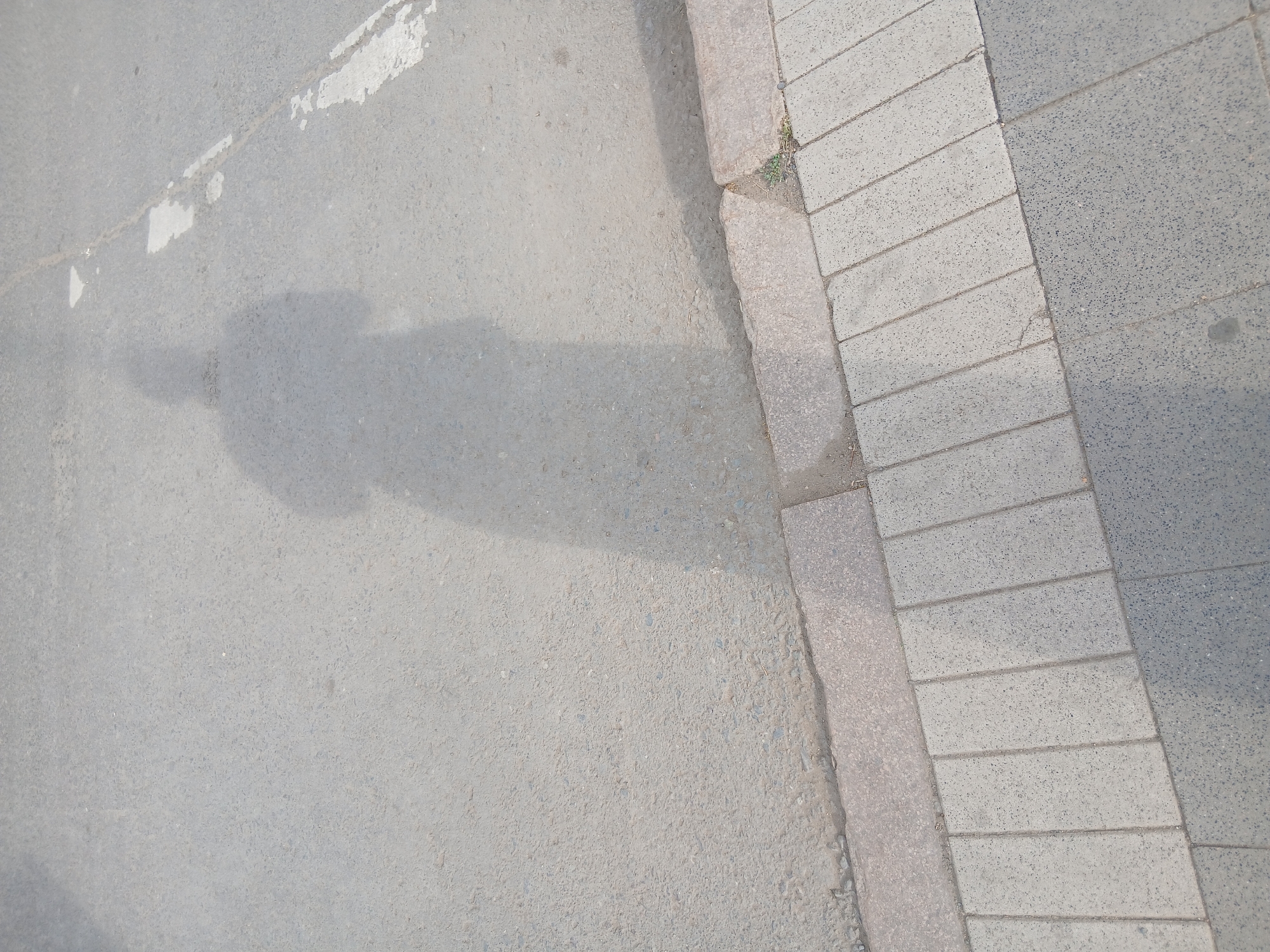 The image shows a shadow of a person on a paved surface, likely a sidewalk or street, in Astana, Kazakhstan. The shadow is cast by the sun from the right, creating a elongated silhouette of an individual wearing what appears to be a long garment. The ground is a mix of asphalt and concrete paving stones, with a curb separating the two. Faint white markings on the asphalt suggest a road or pedestrian crossing. The environment appears to be outdoors during daylight hours, with a focus on the ground and the shadow.