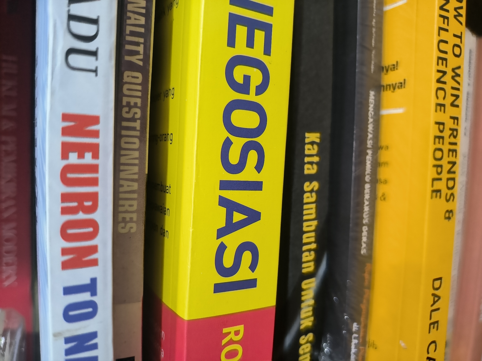 The image shows a close-up of several books stacked together. The book spines are visible, with the following titles and partial titles discernible: "HOW TO WIN FRIENDS & INFLUENCE PEOPLE" by Dale Carnegie, "NEGOSIASI," "NALITY QUESTIONNAIRES," and "NEURON TO N." In addition, there is the phrase "Kata Sambutan Untuk Sem..." visible. The book covers show various color schemes, primarily yellow, red, white, and black. The setting appears to be a bookstore or library. The location is implied to be in the city of Lhokseumawe, Indonesia, due to the presence of text in Bahasa Indonesian.