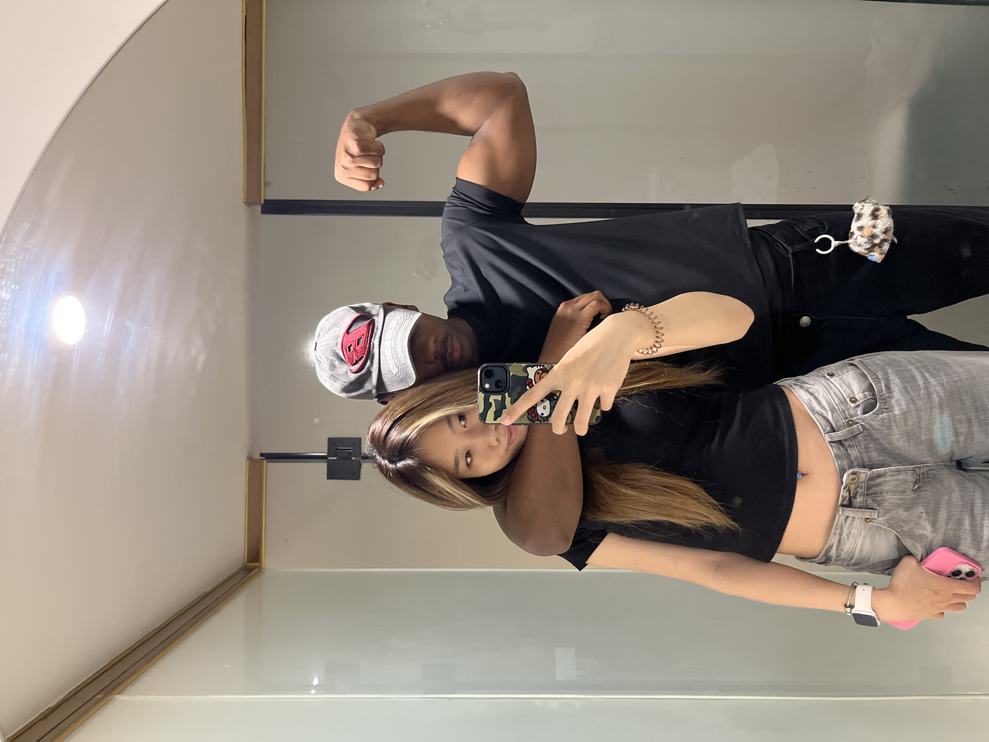 An indoor mirror selfie captures two individuals, a man and a woman, in what appears to be a brightly lit room, possibly a bathroom or changing area in Yuzhong District, China. The image is oriented vertically, but the reflection shows the subjects standing horizontally.
On the left, partially obscured by the woman, is a man with a dark complexion wearing a grey baseball cap with a prominent red, stylized logo that resembles a 'B'. He is dressed in a black t-shirt and is flexing his right bicep towards the camera. A small, leopard-print furry charm with a white tag is clipped to his black pants or belt loop.
On the right, a woman with long, light brown hair with blonde highlights is taking the selfie, holding a smartphone with a camouflage and Hello Kitty-themed case. She looks towards the camera with a slight smile. She wears a black crop top, revealing her midriff and a blue belly button piercing, and light grey jeans. A gold-colored, spiraled bracelet adorns her right wrist, and a white Apple Watch is on her left wrist. She holds a second, pink-cased smartphone in her left hand.
The background shows light-colored walls and reflective surfaces. A bright, circular light fixture is visible in the upper left corner of the reflection, creating a noticeable flare. Golden-hued trim is seen along the bottom and right edges of the mirrored surface, with a black horizontal bar or shelf behind the man. The overall mood is casual and playful.