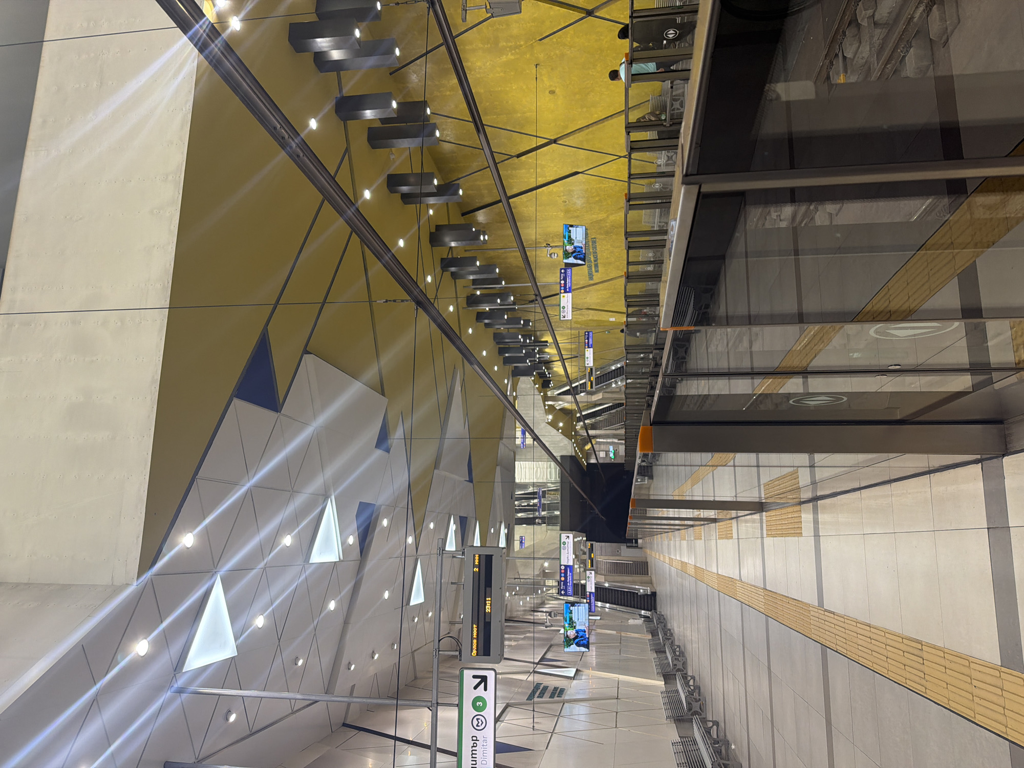 This image depicts the inside of a metro station in Sofia, Bulgaria. The station is characterized by a modern design, featuring geometric patterns and a bright color scheme. The walls on the left side are primarily white with triangular inlays that are illuminated. The ceiling is yellow and includes hanging signs and lighting fixtures. There are tracks and glass barriers to the right side of the image. There are a few people near the upper-right of the image. The floor is gray and includes a tactile paving strip for the visually impaired. Metro signs are visible. The platform has metal benches for seating.