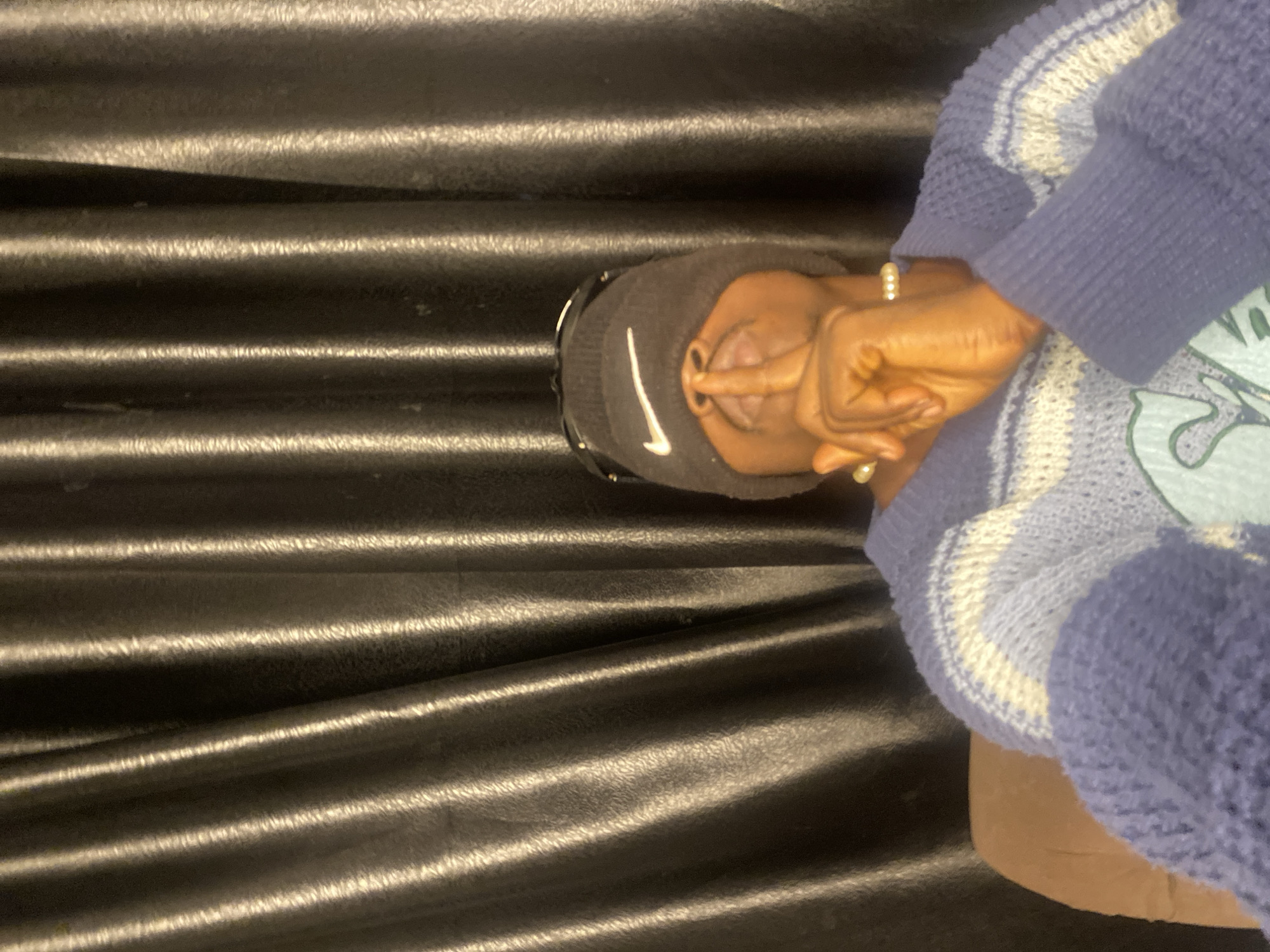 The image shows a close-up of a person from the chest up, with a black curtain-like background. The person is wearing a dark beanie with a white Nike logo and sunglasses pushed up onto the beanie. They are holding a finger to their lips in a shushing gesture, with their eyes closed. A pearl bracelet is visible on their wrist, and they are wearing a light blue and white striped sweater. The lighting is somewhat dim, with highlights on the curtain and the person's skin. There is no discernible location within the city of Abakaliki, Nigeria.