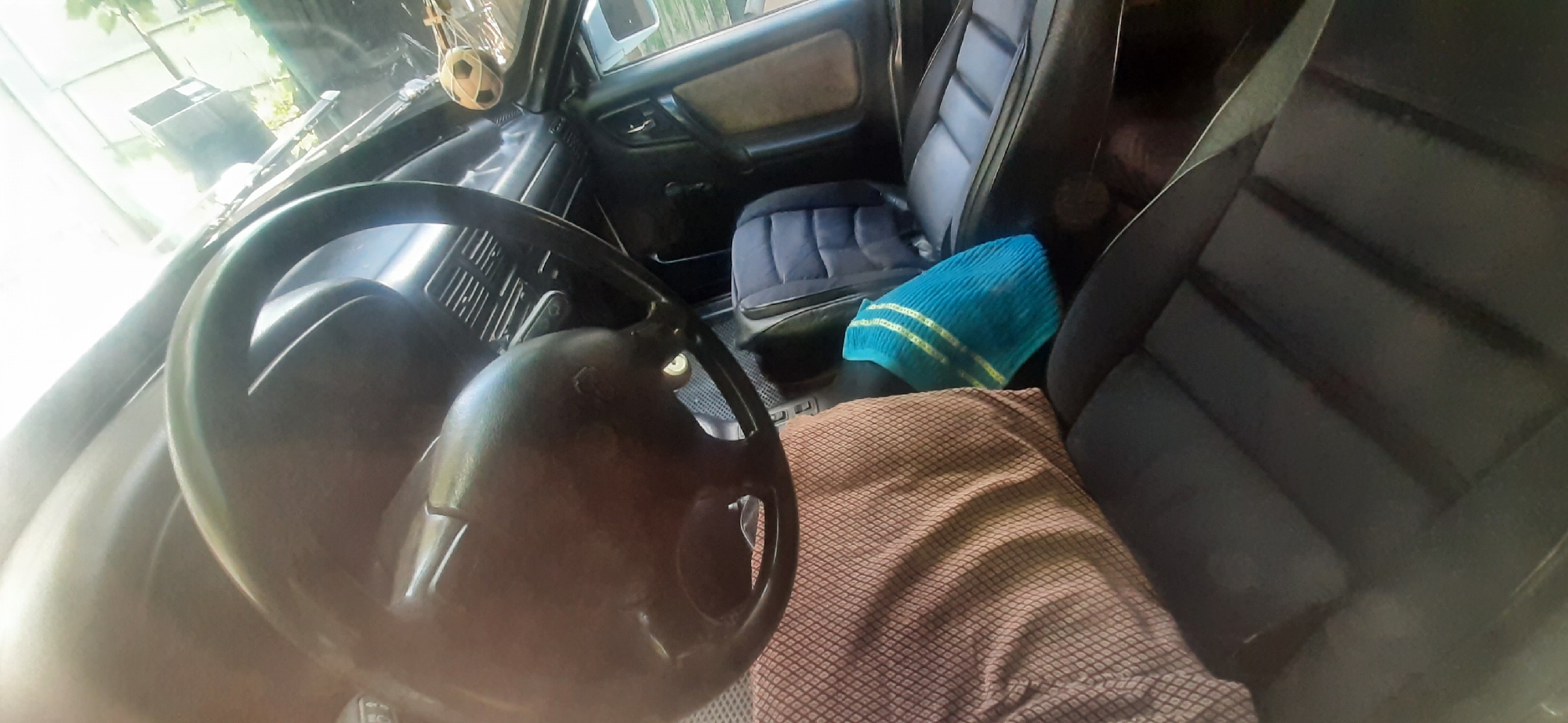 This is a first-person perspective image taken inside a car, likely parked outdoors in Yessentukskaya, Russia. The interior appears to be older, with fabric seats. The driver's seat is covered with a patterned fabric. A blue towel with yellow stripes is placed on the passenger seat. The steering wheel is visible in the foreground. Sunlight streams in from the front windshield, partially obscuring some details. The background suggests an outdoor environment with greenery and structures.