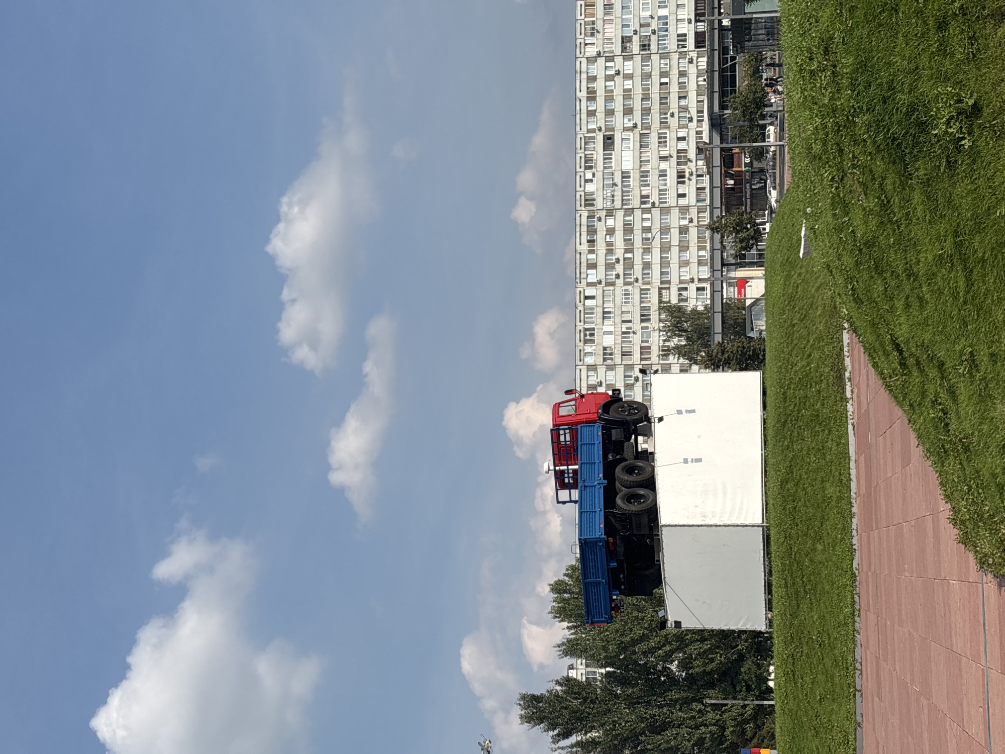 An art installation features a red and blue flatbed truck positioned atop a white, rectangular pedestal, which is itself situated on a grassy mound. The pedestal appears to be covered with a white tarp. Behind the installation, a long, multi-story apartment building with numerous windows dominates the background. To the left of the building and partially obscured by the truck's pedestal are several tall, mature trees. The sky above is blue with scattered white clouds. In the foreground, a red brick pathway is visible, bordered by green grass. The scene suggests an urban outdoor setting in Naberezhnye Chelny, Russia, likely a public space or park area.