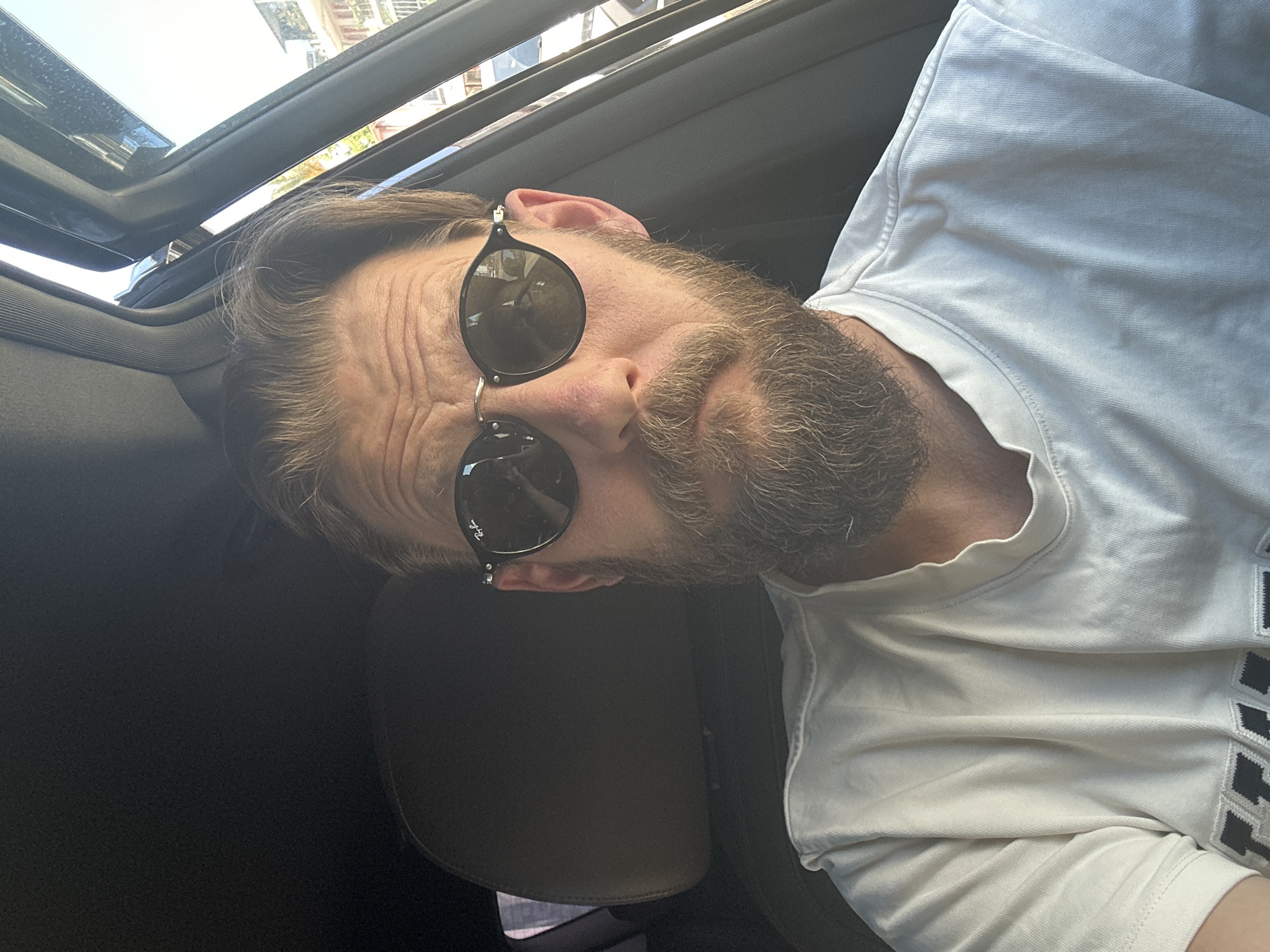 A man with a beard and sunglasses is sitting in a car with an open sunroof. The man is wearing a white t-shirt. The location is possibly Fethiye, Turkey.