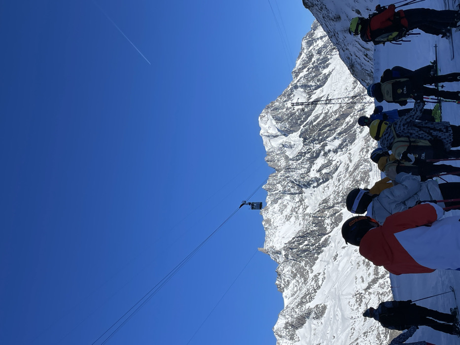 The image, taken on a bright, sunny day, depicts a group of skiers in a high alpine environment, likely Courmayeur, Italy. In the foreground, several individuals, dressed in full winter ski gear including helmets, goggles, and backpacks, stand on skis. Most are facing away from the camera, looking towards the vast, snow-covered mountain range that dominates the right side of the frame. Visible brands on their equipment include "SMITH" and "SALOMON" on helmets, and "CVA" on multiple backpacks.
The sky on the left is a clear, deep blue, with a single white airplane contrail visible in the upper left quadrant. In the mid-ground, a cable car cabin is suspended by cables against the backdrop of the mountains, suggesting a ski lift system. A tall metal structure, possibly a support pylon for the lift, also rises from the snowy slopes. The mountains are rugged, characterized by dramatic rocky outcrops and expansive snowfields, reflecting the bright sunlight.
The scene captures a moment of anticipation or gathering, possibly before a descent or while waiting for a lift. The clear weather and pristine snow suggest ideal conditions for skiing or snowboarding, amidst a breathtaking natural landscape.