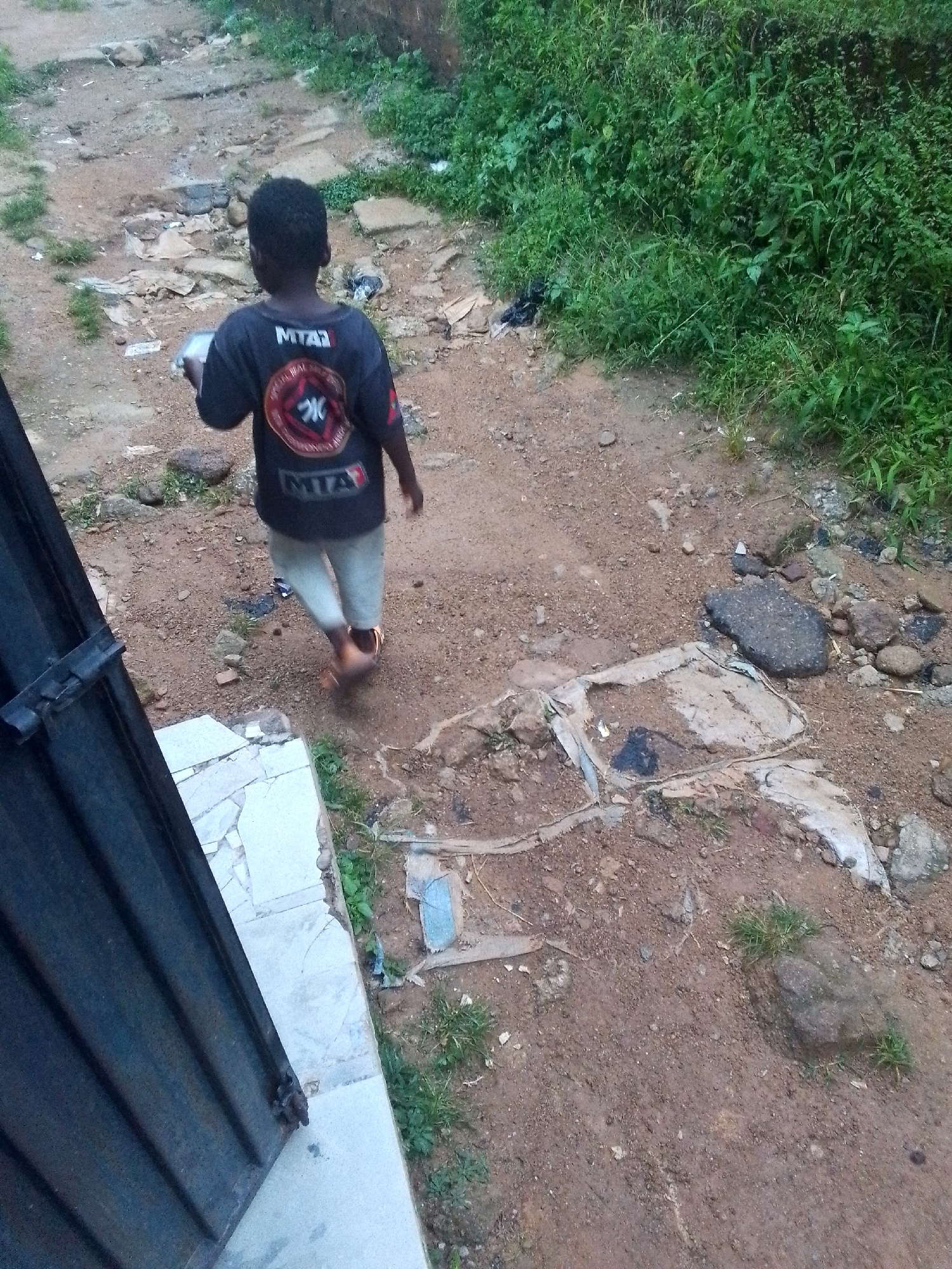 The image depicts a young boy walking away from the camera on a dirt path in Jos, Nigeria. He is wearing a dark t-shirt with a circular logo on the back and light-colored trousers. The boy is carrying a white rectangular object in his right hand. The path is uneven and appears to be in a semi-urban or rural area, with patches of green vegetation on either side. Debris, including what appears to be discarded plastic and rocks, is scattered along the path. A portion of a dark, corrugated metal structure, possibly a gate or fence, is visible on the left side of the frame. The overall environment suggests a common outdoor setting within the city, characterized by unpaved surfaces and natural growth.