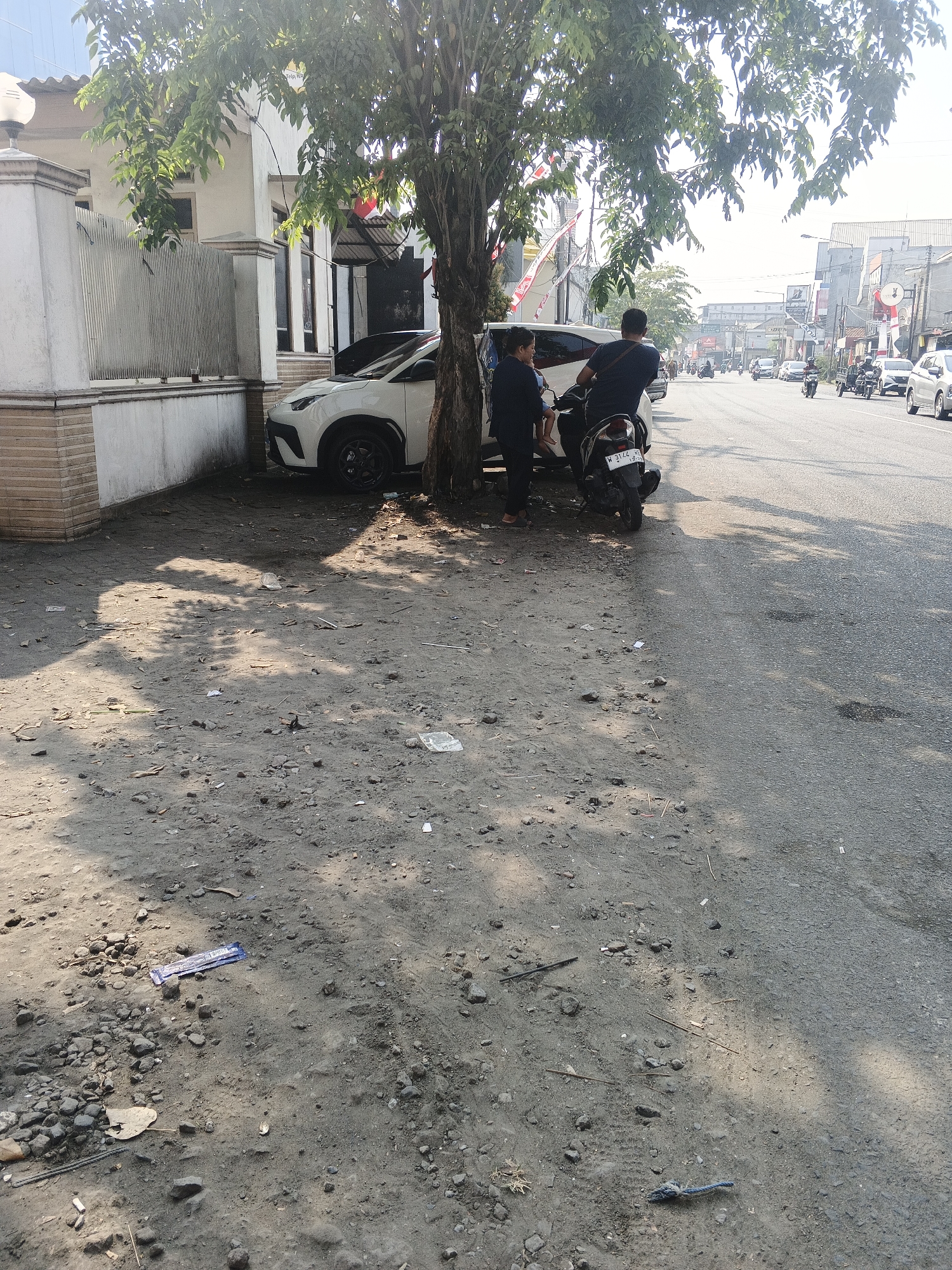 In Kemangsen, Indonesia, an outdoor scene captures a street with a paved roadway and a dirt shoulder. On the dirt shoulder, a white car is parked beside a tree with green foliage. Two individuals are present: one is standing and the other is seated on a black scooter parked near the car. The scooter has a license plate visible. The street is lined with buildings and other vehicles, including motorbikes and cars, suggesting a moderately busy urban environment. Red and white flags are displayed on some of the buildings, possibly indicating a national celebration. The ground on the shoulder is littered with debris, including small rocks and scattered papers. Sunlight casts shadows from the tree and overhead elements onto the ground.