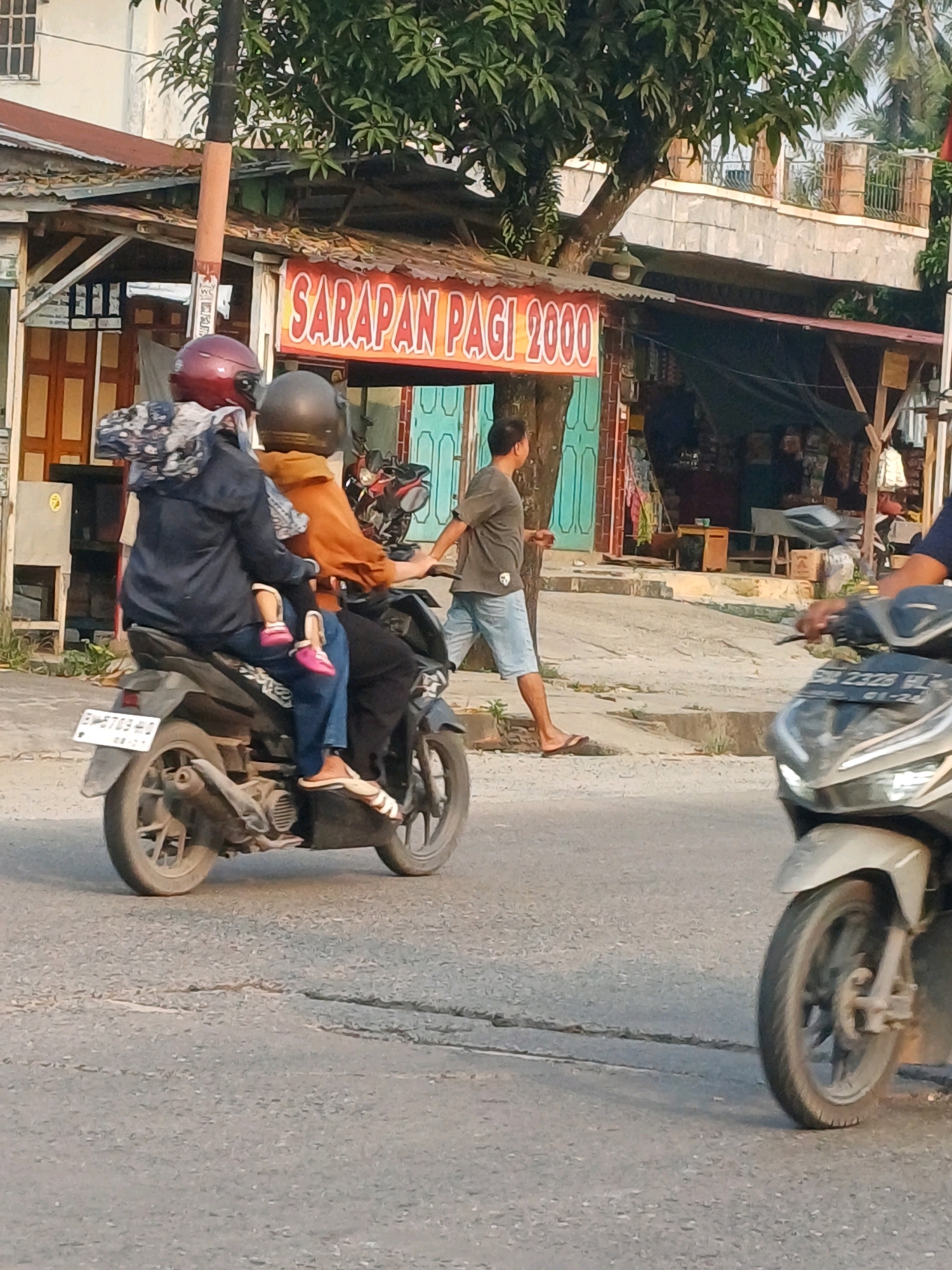 The image shows a street scene in Dumai City, Indonesia. In the foreground, a motorcycle with three passengers is moving from left to right. The driver wears a dark jacket and a red helmet. Behind the driver is a person wearing a brown shirt and a black helmet, holding a child. The child, wearing pink shoes, is seated between the two adults. The motorcycle has a license plate with the inscription "B 6703 EA". A second motorcycle, partially visible on the right side of the image, is also moving from left to right. In the background, a building with a red awning displaying the text "SARAPAN PAGI 2000" is visible. A man in a gray t-shirt and blue shorts is walking on the sidewalk to the left of the motorcycle. Trees and other buildings are also present in the background. The road surface appears to be asphalt with some cracks.