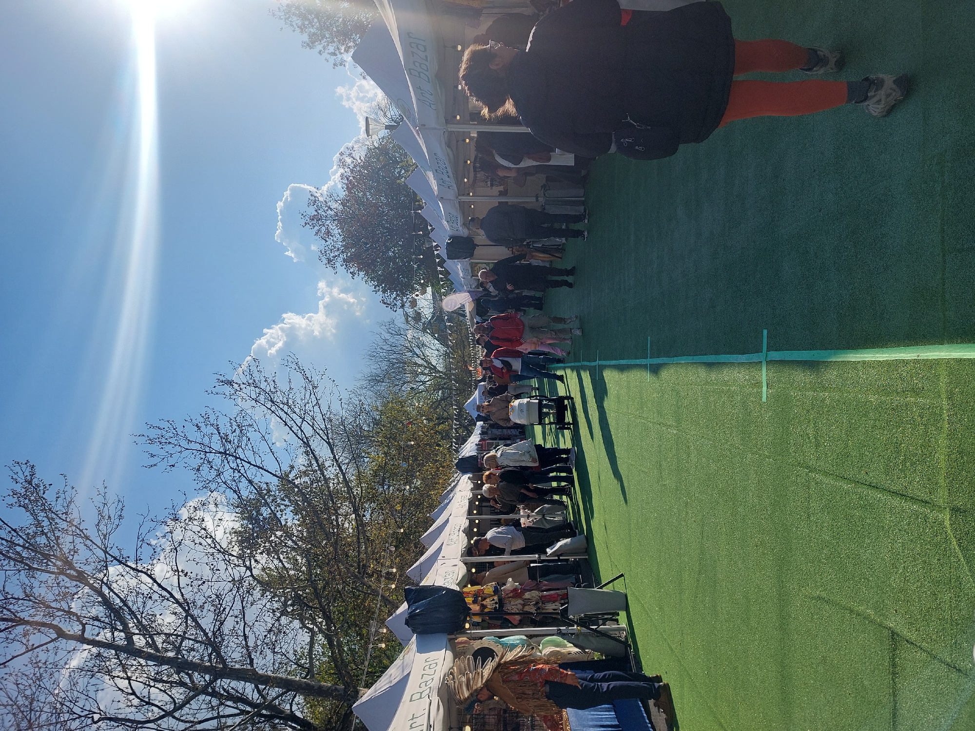 An outdoor market or bazaar is depicted on a bright, sunny day with a clear blue sky and fluffy white clouds. The sun shines intensely from the upper left, casting a strong lens flare across the image. The ground is covered in vibrant green artificial turf, marked with faint white lines, suggesting a repurposed sports court.
Numerous people are visible, casually dressed, walking and browsing along a row of white canopy tents that line the market. Several tents clearly display the text "Art Bazar". In the right foreground, a person wearing a black jacket and bright orange pants stands with their back to the viewer, facing deeper into the market area. Further down the row of stalls, other attendees are seen looking at the merchandise.
Bare-branched trees are visible on the left side of the frame, with some autumnal foliage on trees to the right, suggesting a setting in late autumn or early spring. The atmosphere appears lively and bustling, indicating an active public event.