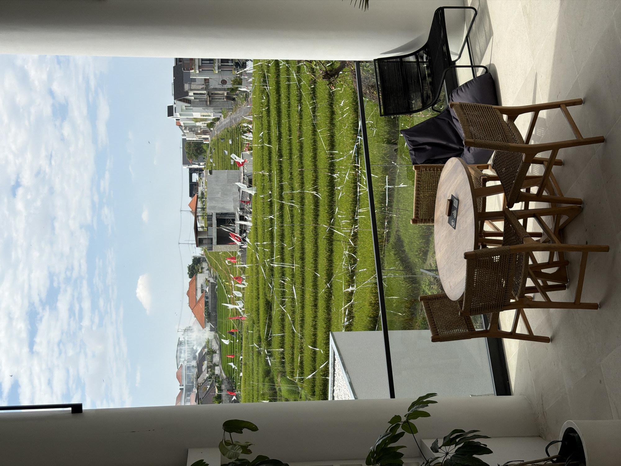 The image depicts an outdoor seating area with a view of terraced rice fields under a partially cloudy sky. In the foreground, a round wooden table is surrounded by four woven chairs. To the right of the table, a dark beanbag chair and a black wire-framed chair are visible. A potted plant is situated to the left of the seating area. The background showcases vibrant green rice paddies, with numerous white and red flags or banners strung across them. Several buildings, some appearing under construction, are visible in the distance, along with a road winding through the landscape. Smoke is emanating from one of the structures. The overall impression is that of a scenic location, possibly a cafe or restaurant with an agricultural backdrop.